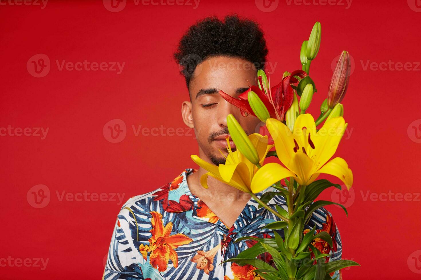 Portrait of young calming African American man, wears in Hawaiian shirt, holds yellow and red flowers bouquet and covered face, stands over red background with closed eyes. photo