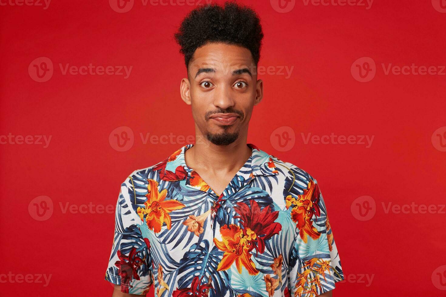 joven confuso africano americano chico usa en hawaiano camisa, mirando a el cámara con preguntado y malentendido, soportes terminado rojo antecedentes con cruzado brazos. foto