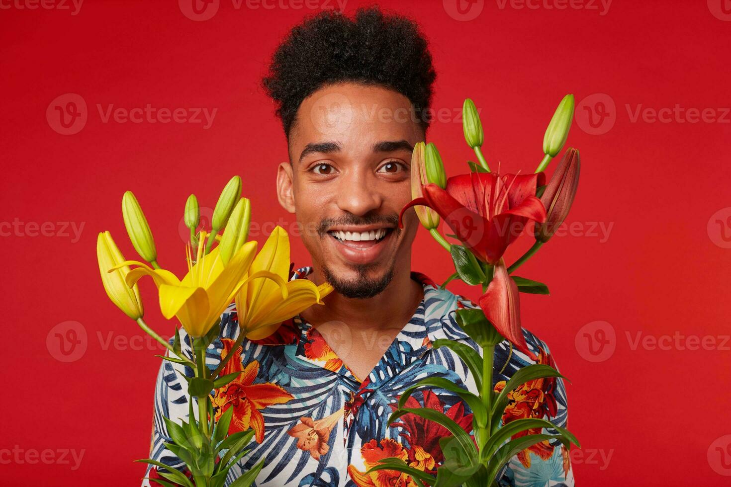 Close up cheerful young African American guy, wears in Hawaiian shirt, looks at the camera with happy expression, holds yellow and red flowers, stands over red background. photo
