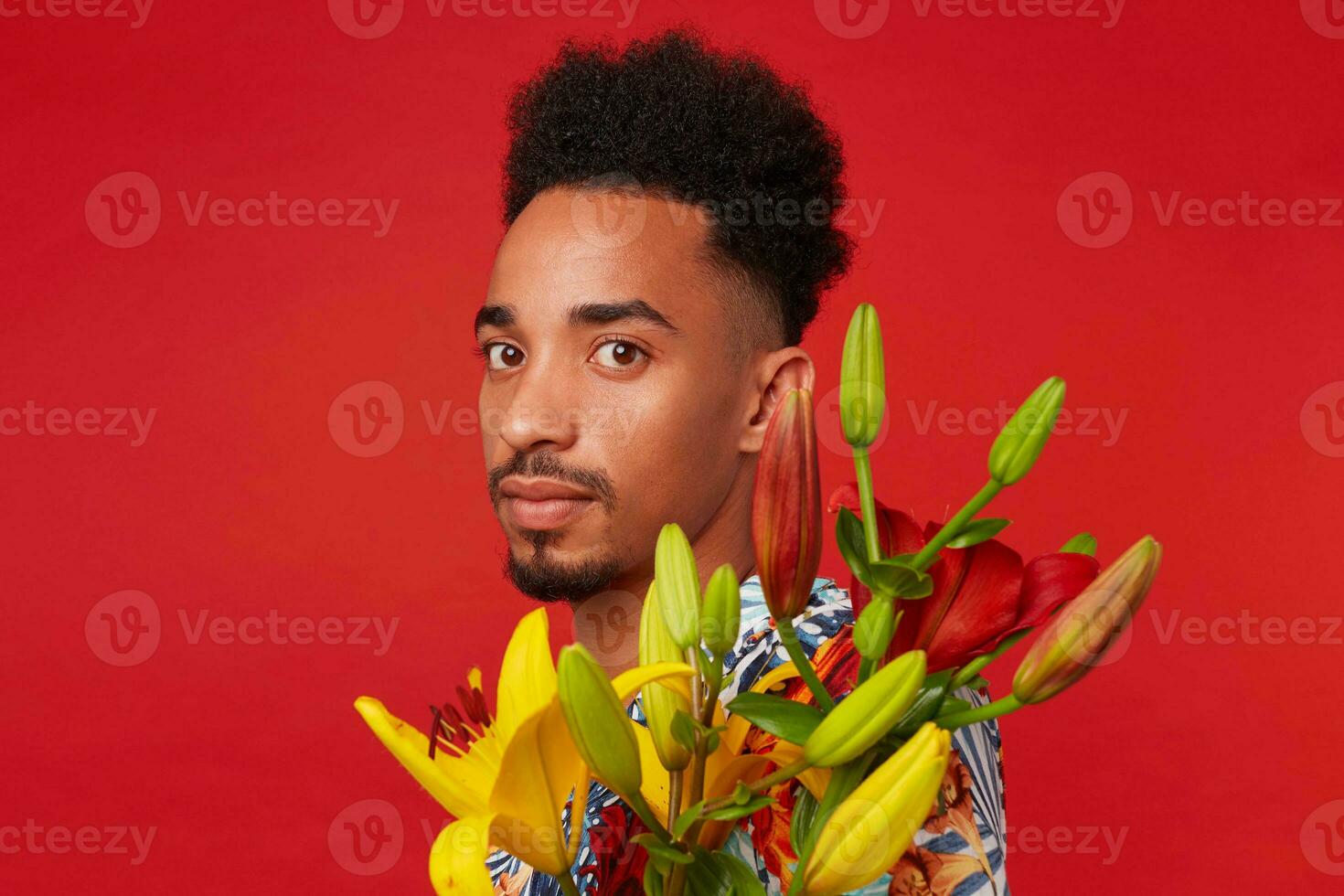 Close up of young calm dark skinned man, wears in Hawaiian shirt, holds yellow and red flowers bouquet, stands over red background. photo