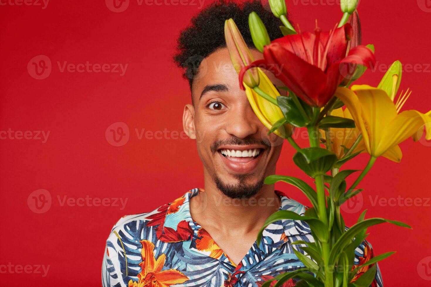 Close up of young laughing African American man in Hawaiian shirt, looks at the camera with happy expression, holds yellow and red flowers bouquet, stands over red background. photo