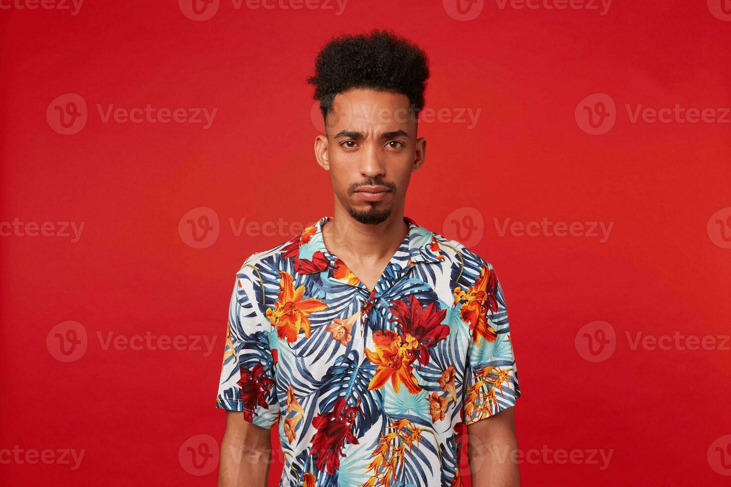 Portrait of frowning young African American guy, wears in Hawaiian shirt, looks at the camera with unhappy expression, stands over red background. photo