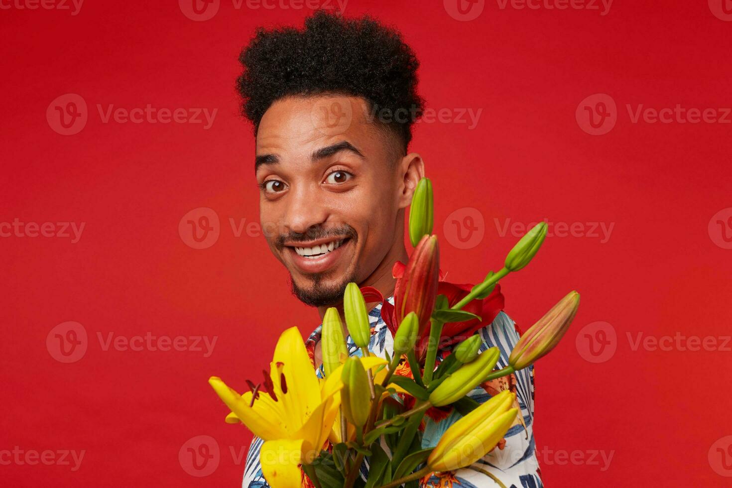 cerca arriba joven positivo oscuro desollado hombre, usa en hawaiano camisa, mira a el cámara con contento expresión, sostiene amarillo y rojo flores, soportes terminado rojo antecedentes y sonriente. foto