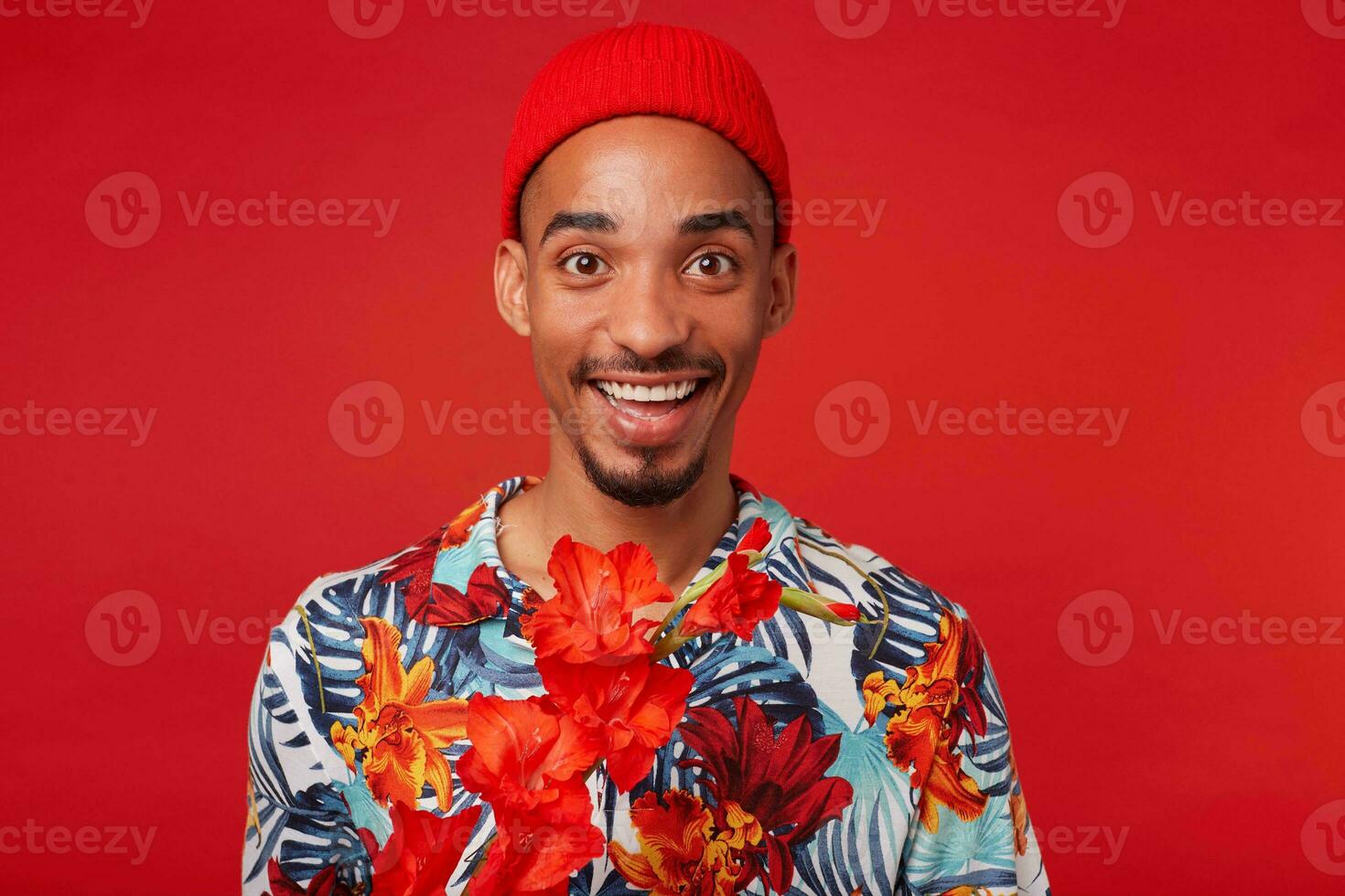 retrato de joven contento oscuro desollado chico, usa en hawaiano camisa y rojo sombrero, mira a el cámara con contento expresión, soportes terminado rojo antecedentes. foto