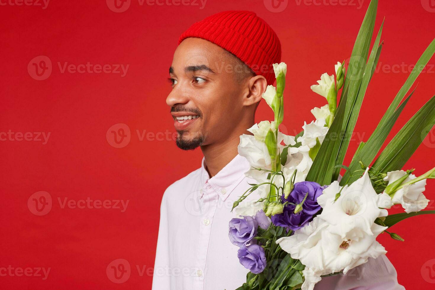 Close up of young positive dark skinned man, wears in white shirt and red hat, stands over red background and broadly smiling. photo