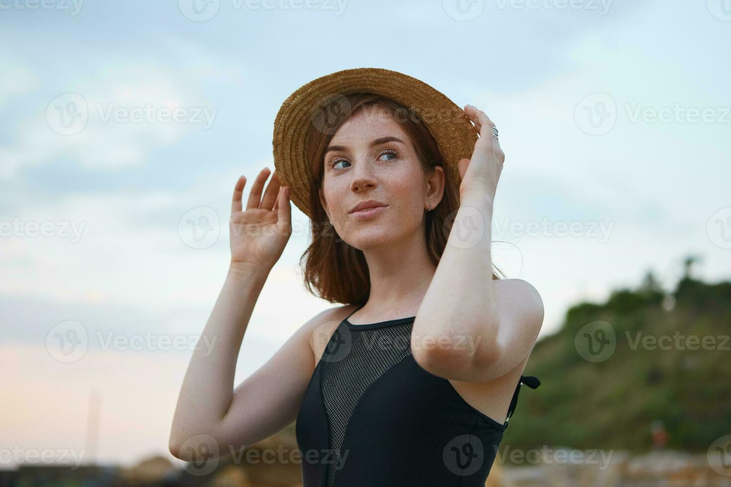 Young ginger cute lady with freckles walks on the beach, holding hat, dreamily looks at the camera, looks positive and happy. photo