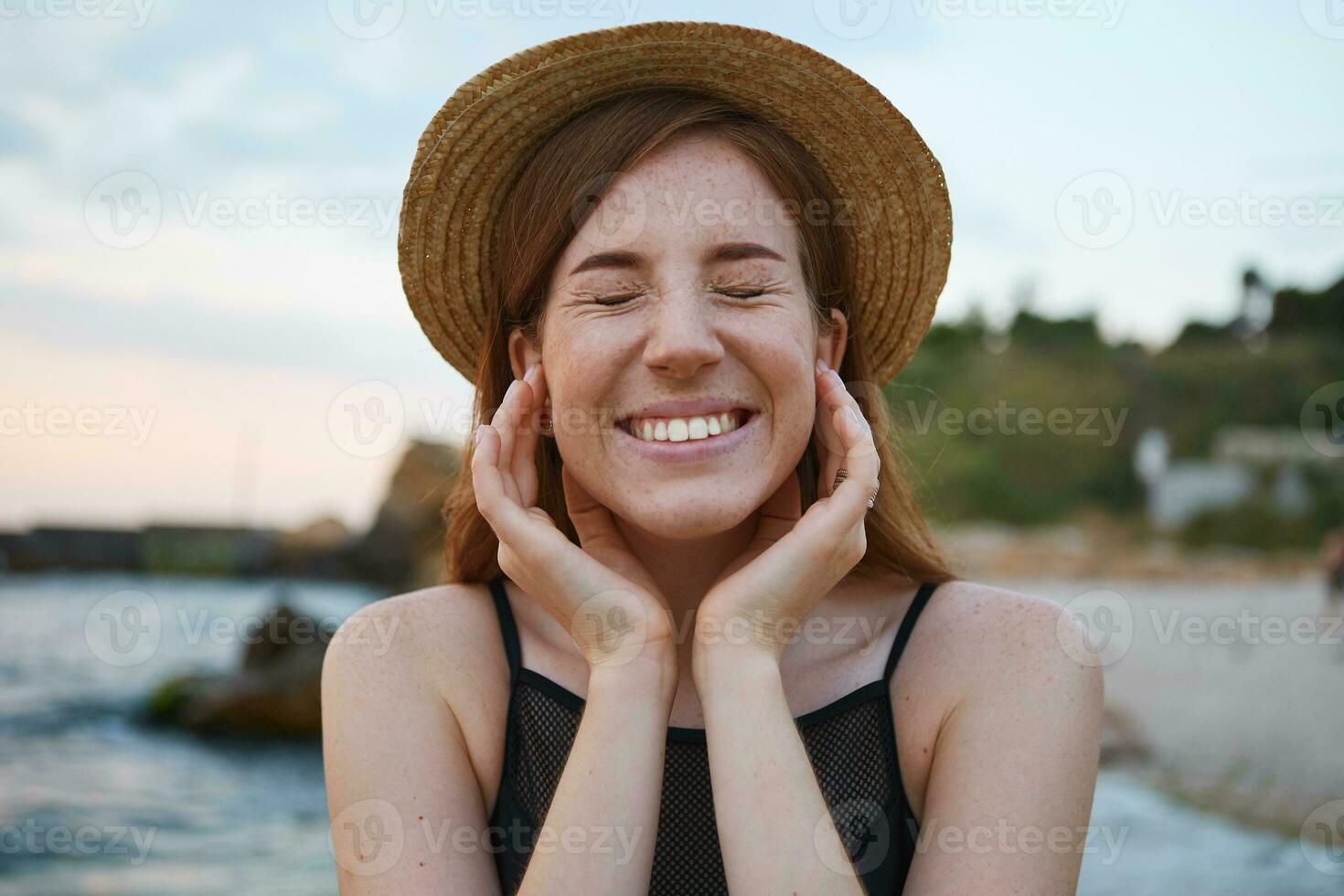 Close up of young ginger cute freckles woman walks on the beach, wears hat, touches cheeks, broadly smiles with closed eyes, looks positive and happy. photo