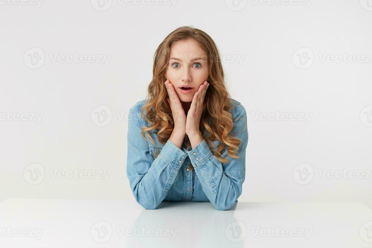 retrato de joven linda preguntado Rizado rubia dama en mezclilla camisas sentado a el blanco mesa y sonrisa, mira sorprendido, aislado terminado blanco antecedentes. foto