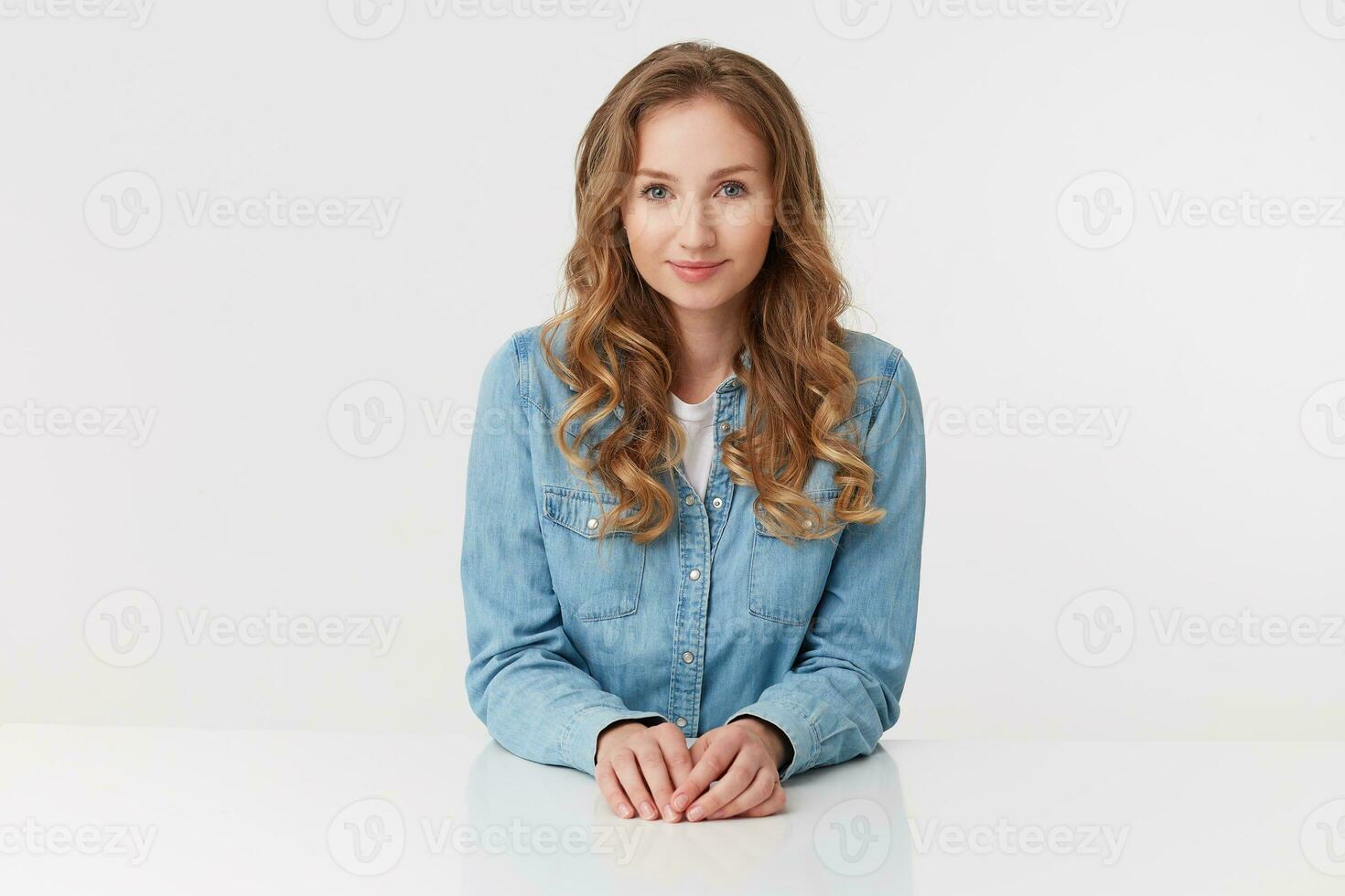 Portrait of young cute curly blonde lady in denim shirts sitting at the white table and smile, looks positive, isolated over white background. photo