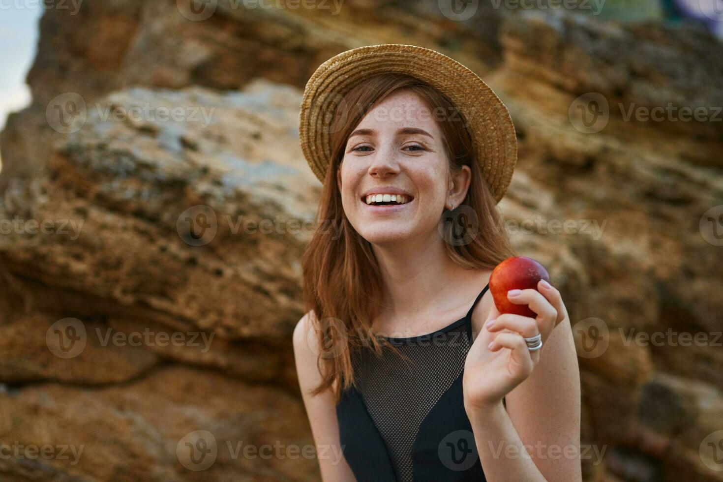 Portrait of young ginger nice freckles lady on the beach, wears hat, eating a peach, broadly smiles and looks at the camera, looks positive and happy. photo