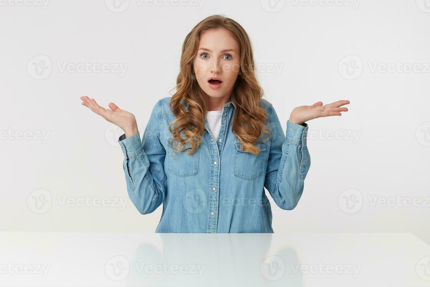 Portrait of young wondered cute curly blonde lady in denim shirts sitting at the white table and spreads his arms to the side, isolated over white background. photo