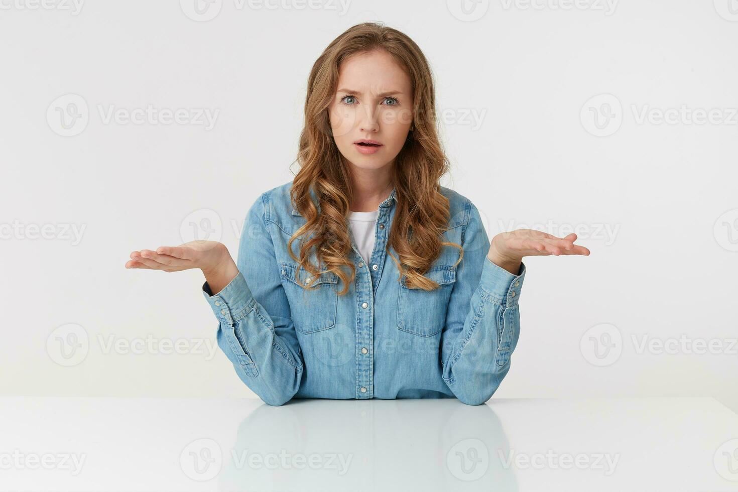Young outraged blonde lady in denim shirts, sitting at the white table and spreads his arms to the side, frowning and looks discontented, isolated over white wall. photo