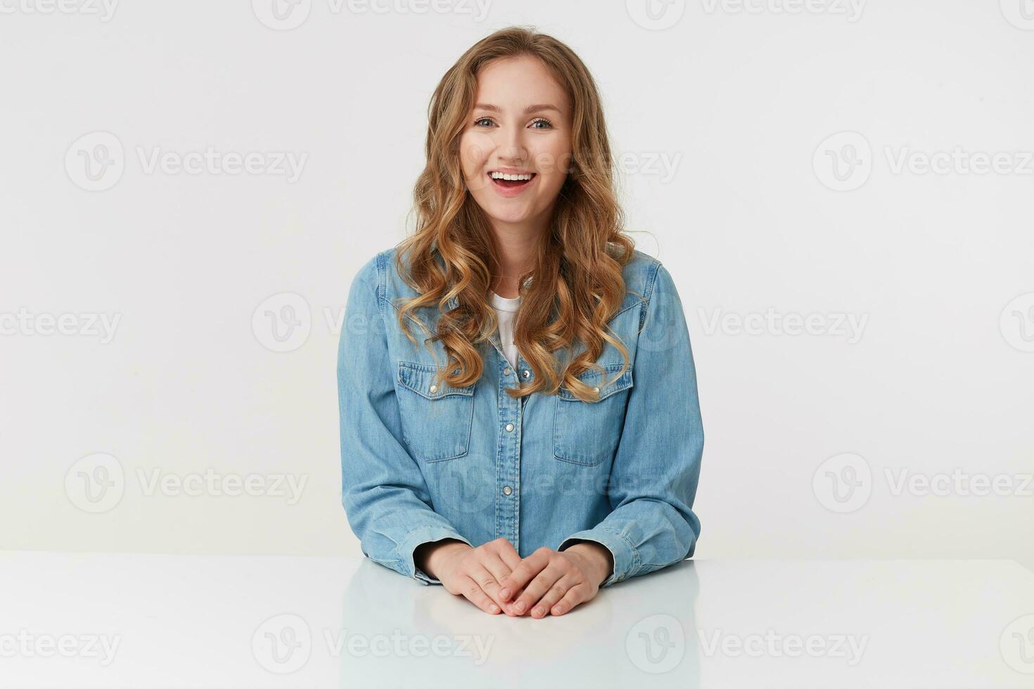 Portrait of young happy cute curly blonde lady in denim shirts sitting at the white table and broadly smiling, isolated over white background. photo