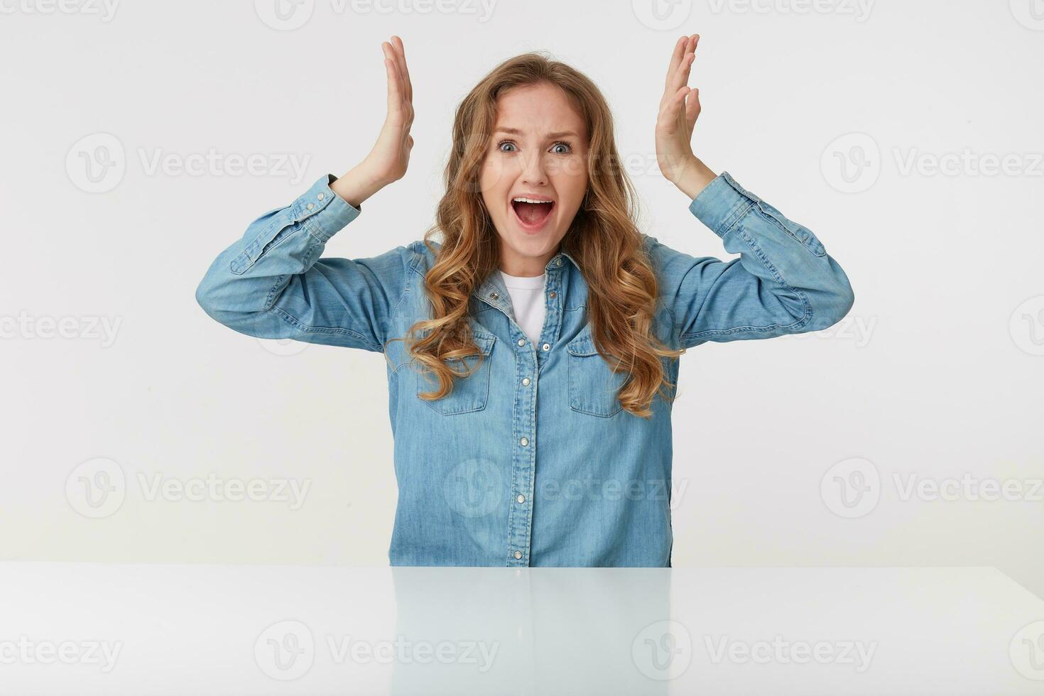 Photo of young aggressive blonde lady wears in denim shirt, screaming and looks angry with raised hands, sitting at the white table over white background.