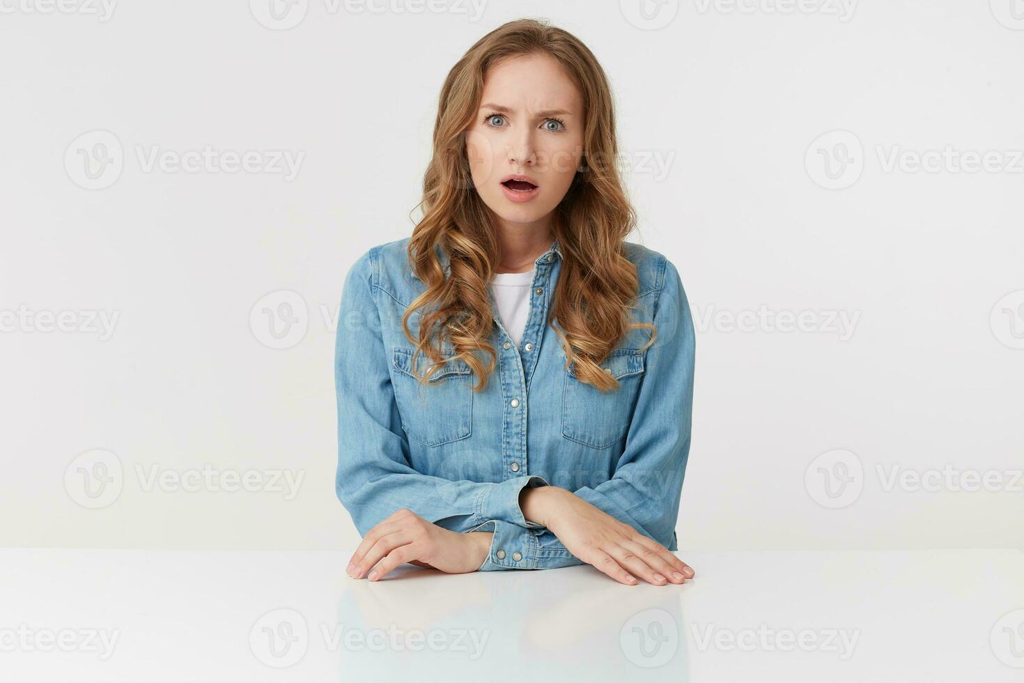 Photo of young outraged blonde woman wears in denim shirts, sitting at the white table, frowning and looks discontented, isolated over white background.