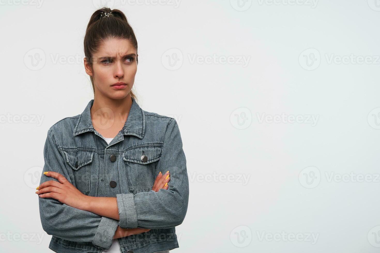 Portrait of young frown brunette lady wears in white t-shirt and denim jackets, looks away with disgusted expression, stands over white background with crossed arms. photo