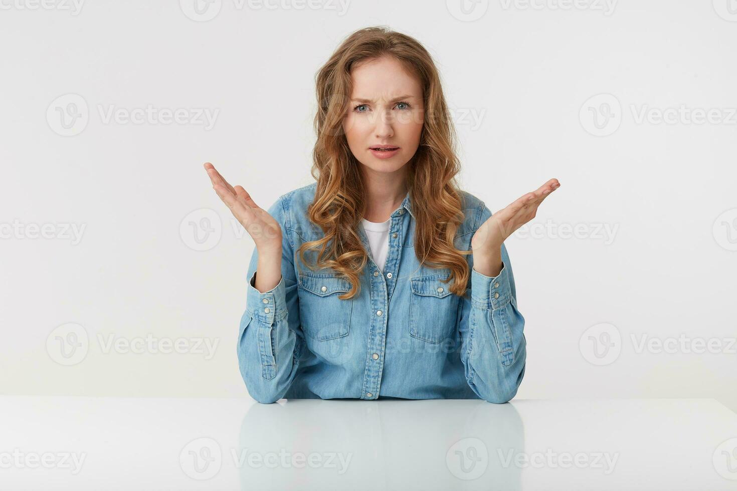 Young outraged blonde lady wears in denim shirts, sitting at the white table and spreads his arms to the side, frowning and looks discontented, isolated over white background. photo