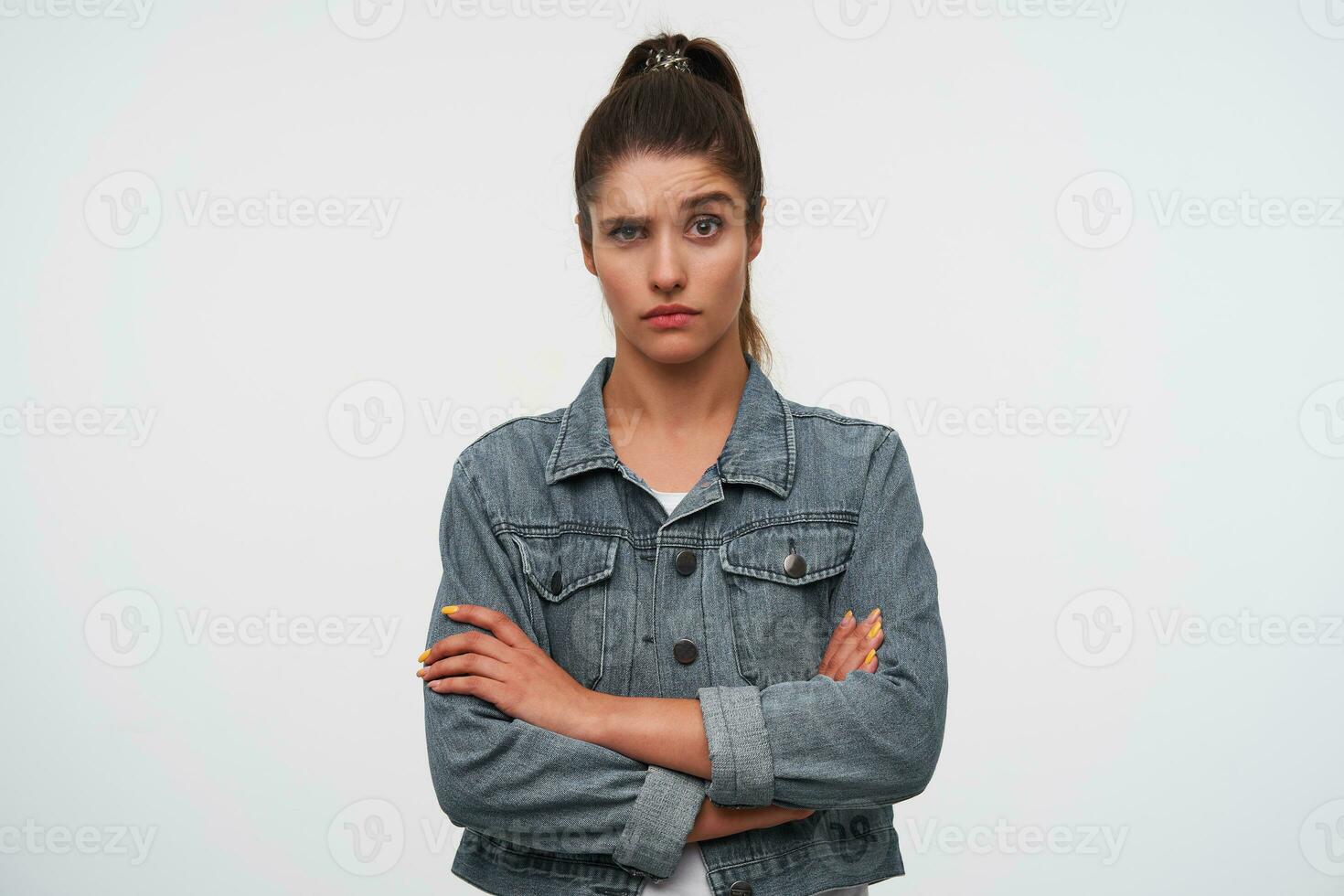 Portrait of young brunette lady with raised eyebrow wears in white t-shirt and denim jackets, looks at the camera with unhappy expression, stands over white background with crossed arms. photo