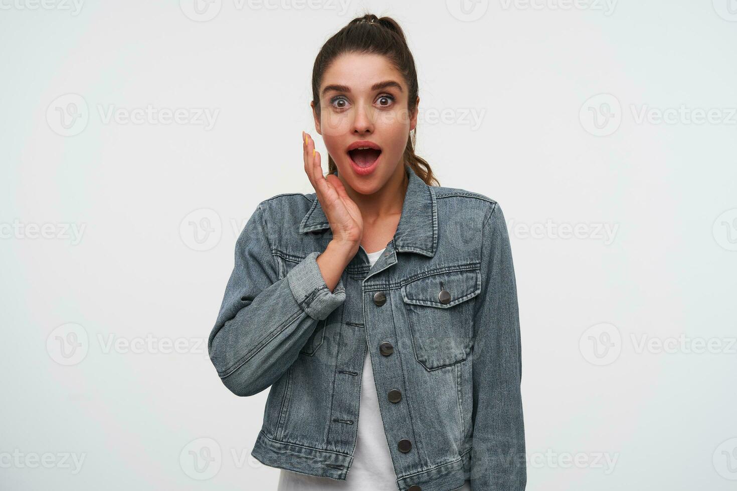 Young happy amazed brunette lady in white t-shirt and denim jackets, looks at the camera with wide open mouth and eyes in surprised expression, keep palm on the cheek, stands over white background. photo