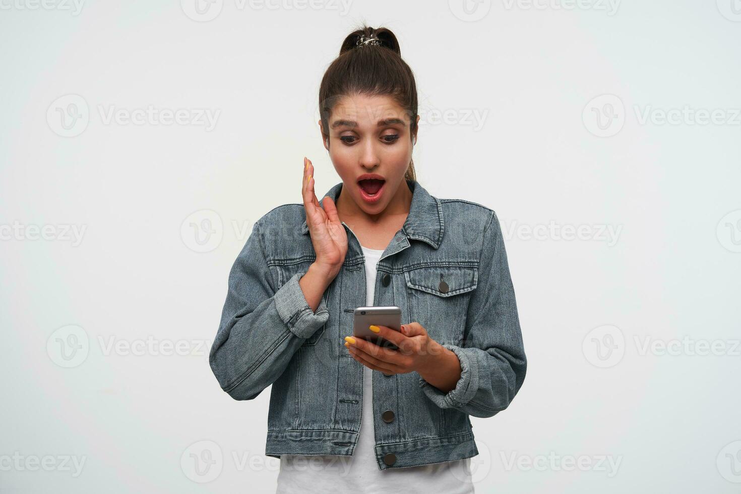 Young shocked brunette lady with wide open mouth in white t-shirt and denim jackets, holds smartphone and looks surprised, listening new song of favorite band. photo