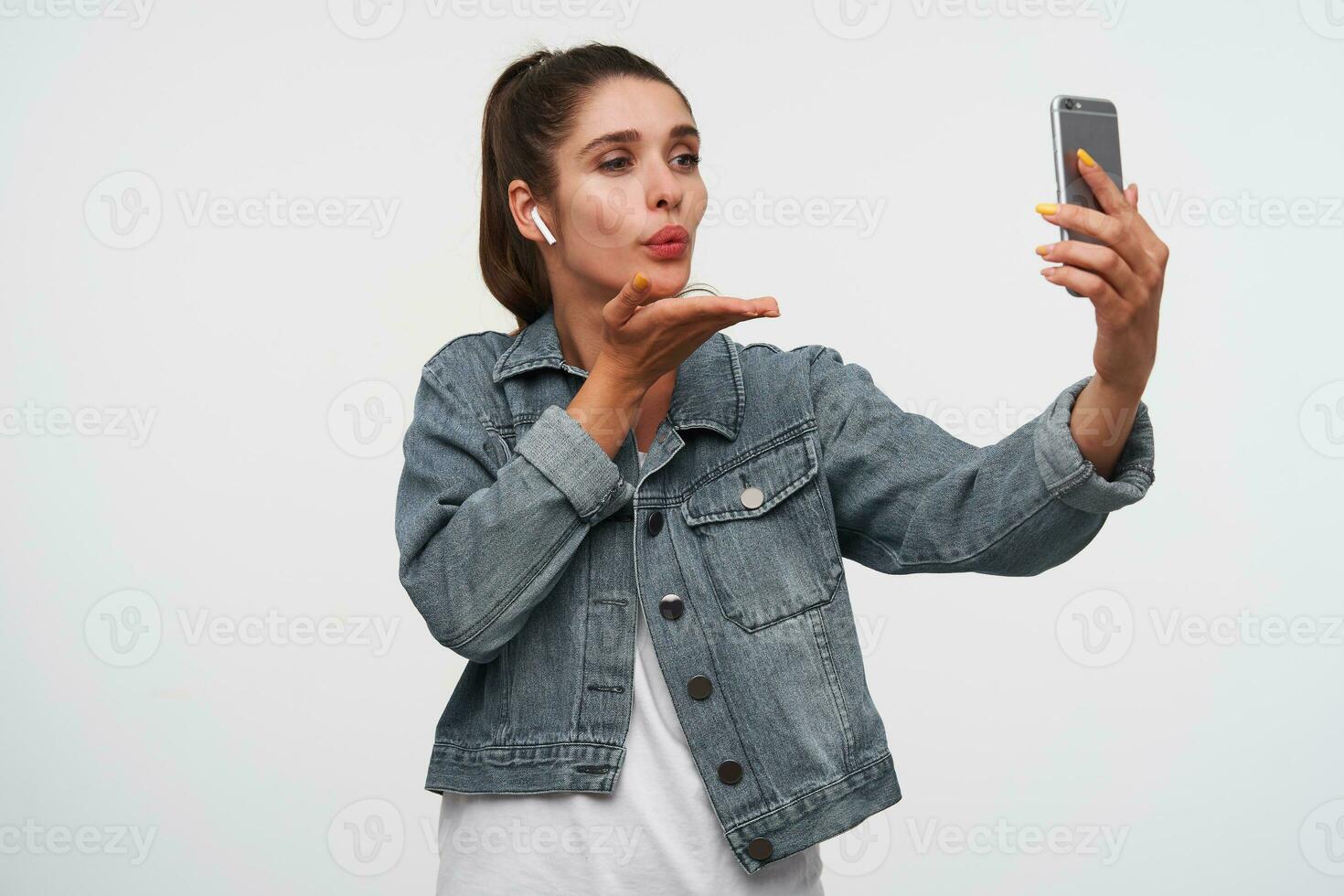 Young happy brunette woman wears in white t-shirt and denim jackets, holds smartphone and sends kiss to the videochat. Stands over white background. photo