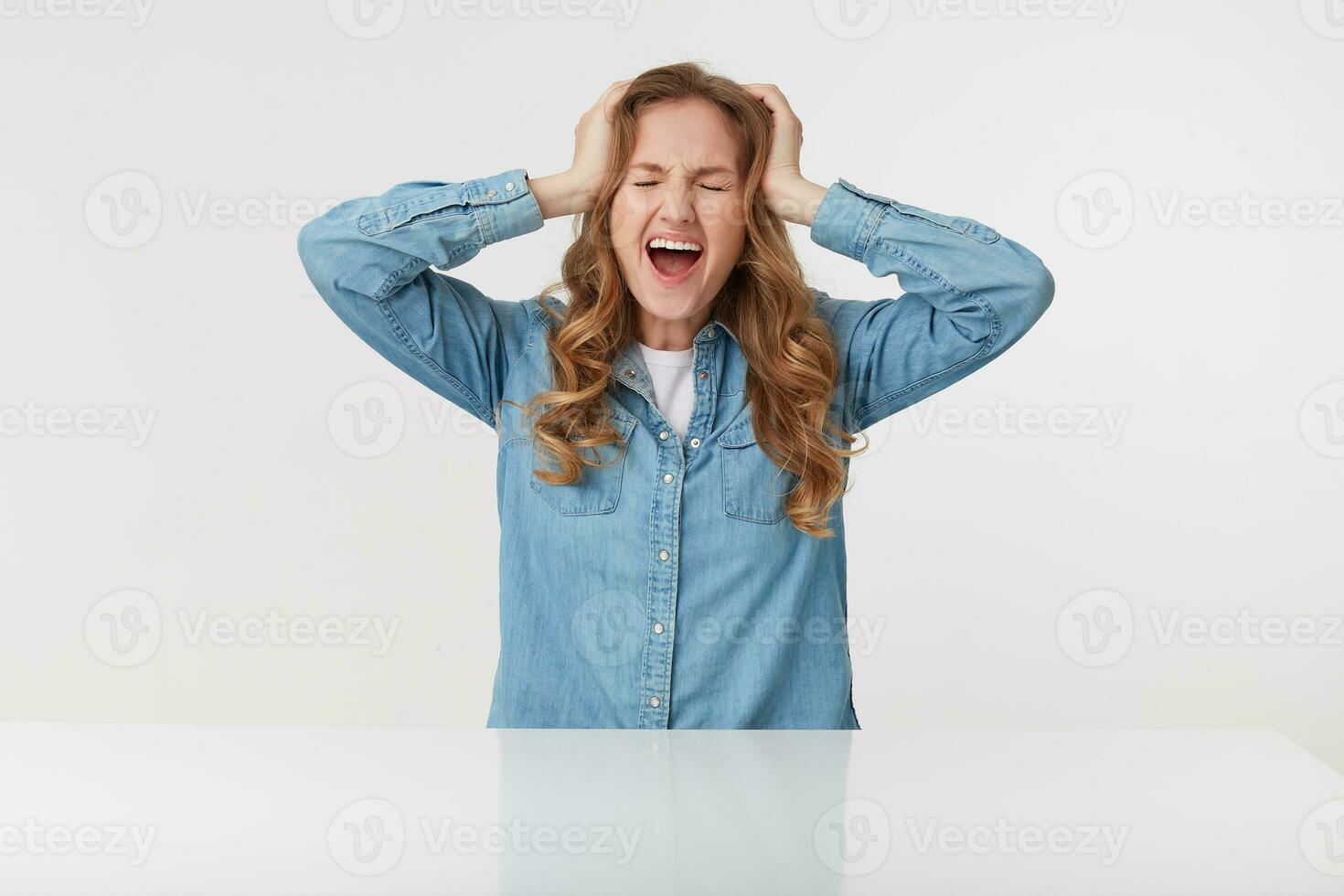 Young shocked blonde lady wears in denim shirt sitting at the white table, screaming with wide open mouth, keep palms on the head, stands over white background. photo