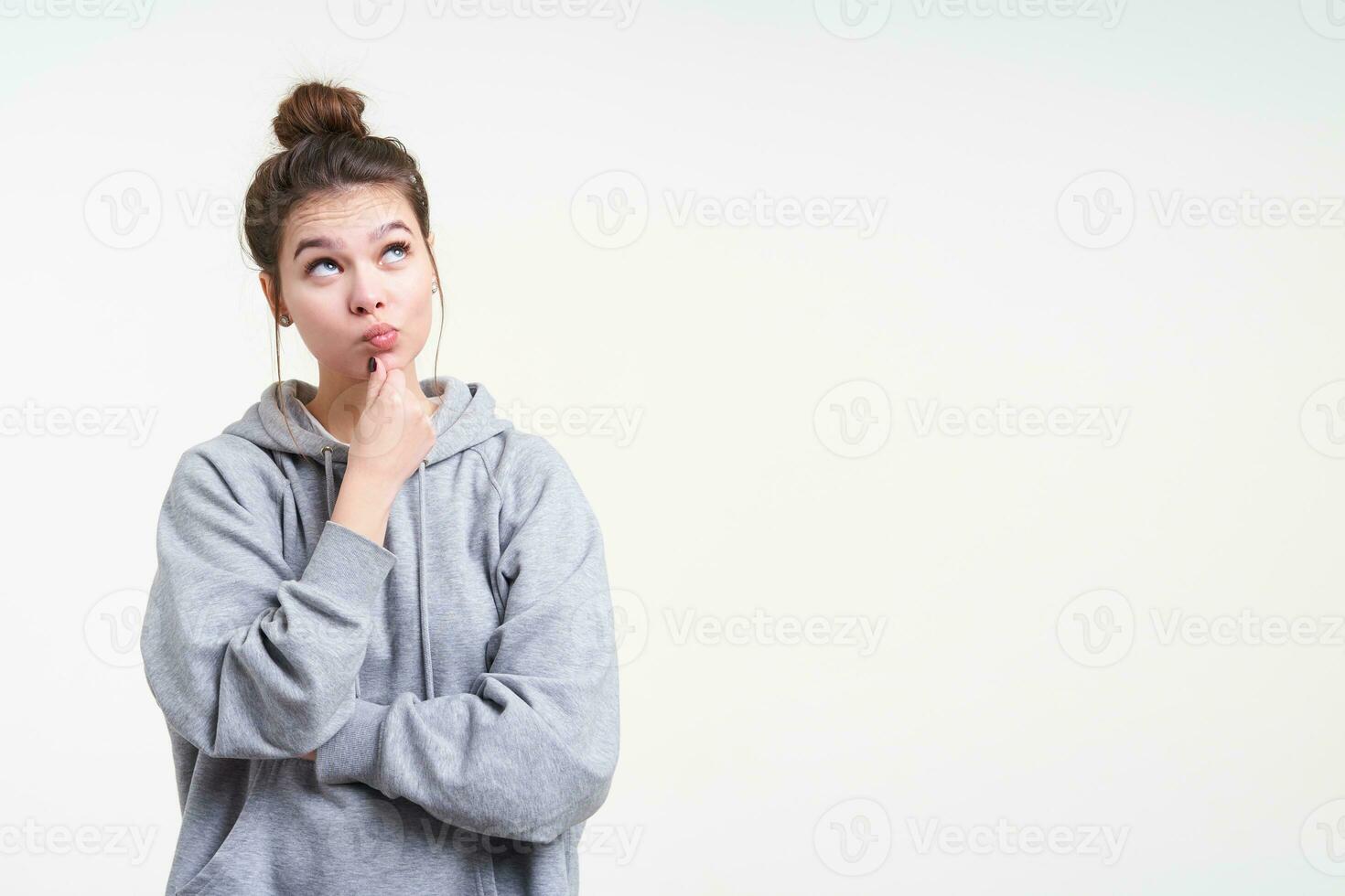 Pensive young attractive brunette female with casual hairstyle holding her chin with raised hand while looking thoughtfully upwards, isolated over white background photo