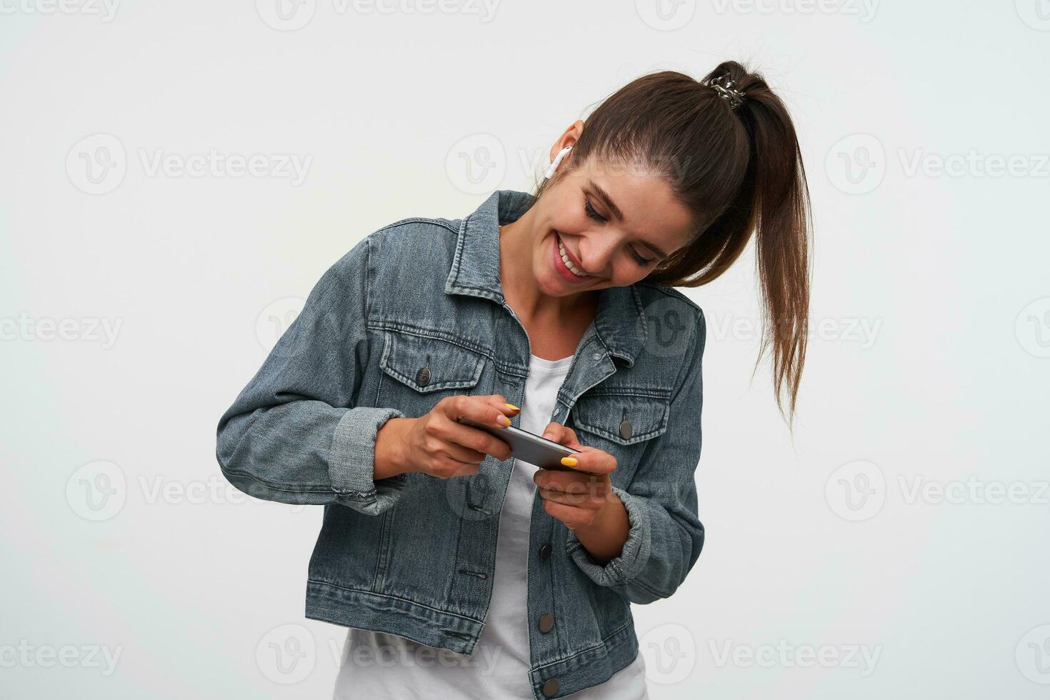 Portrait if young cheerful brunette woman wears in white t-shirt and denim jackets, holds smartphone and sends funny massege to the chat. Stands over white background. photo