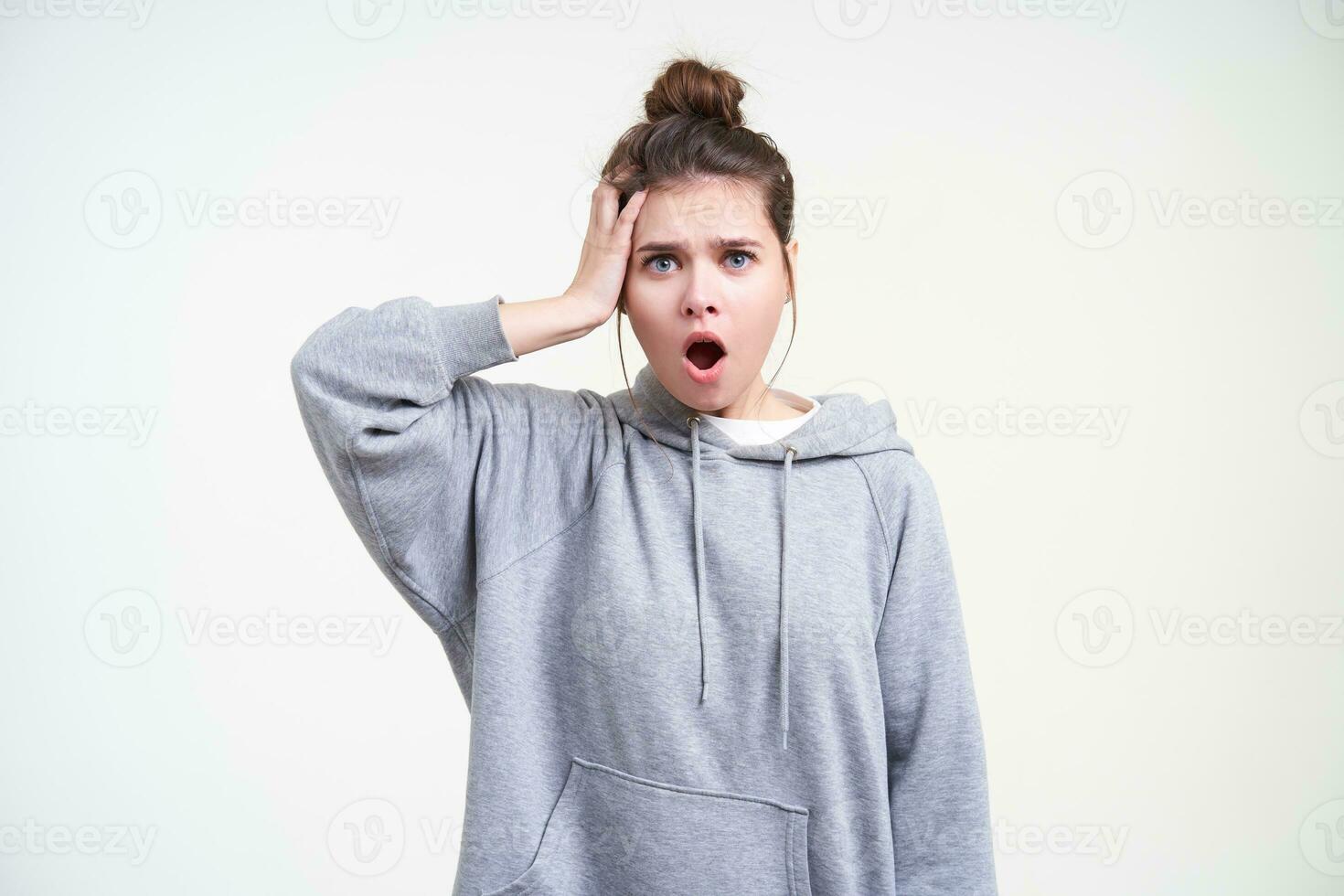 Bewildered young attractive brunette female with bun hairstyle keeping raised palm on her head and looking bemusedly at camera, posing over white background photo