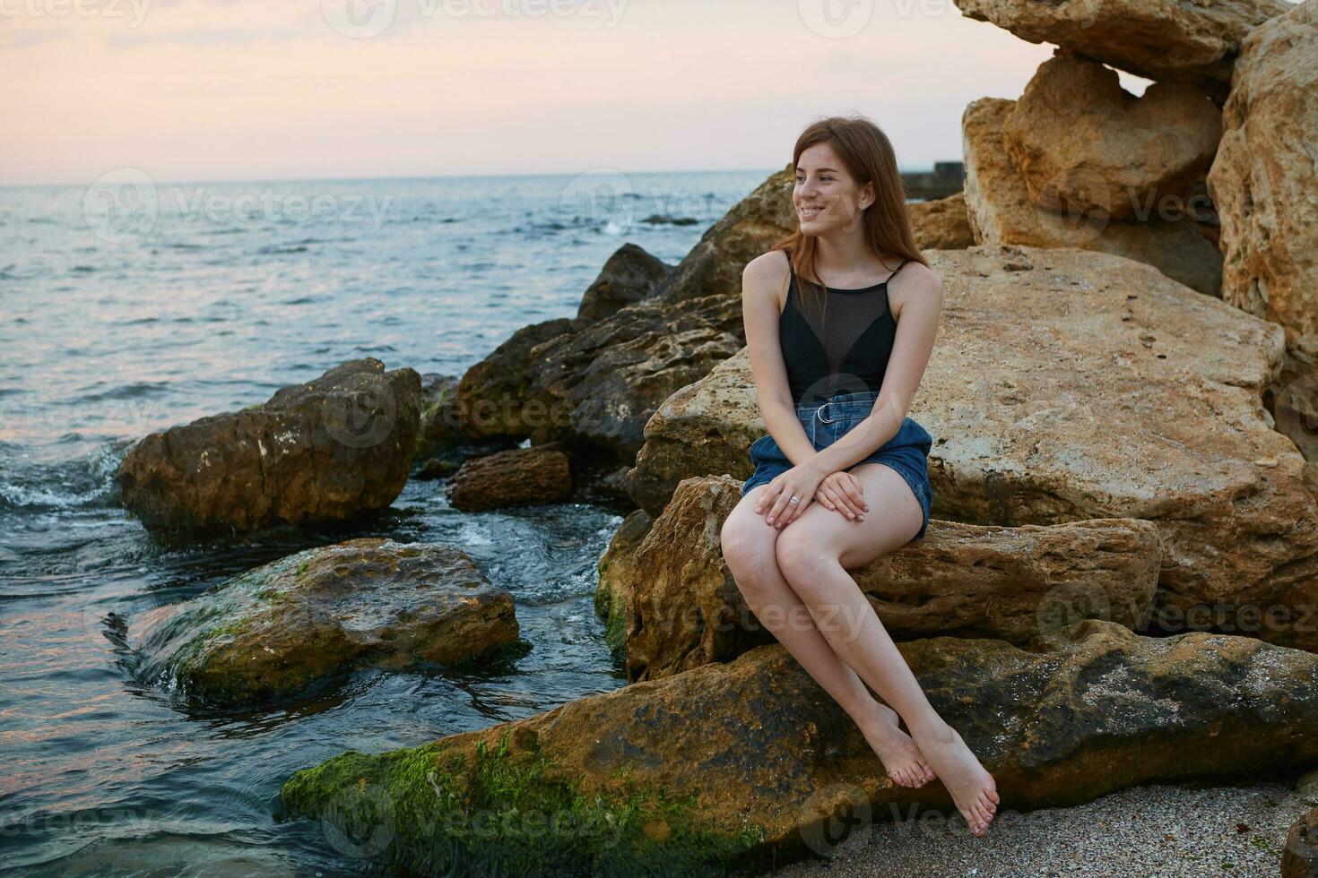 Portrait of young cheerful ginger cute woman with freckles sits on the beach, dreamily looks away and smiles, looks positive and happy. photo