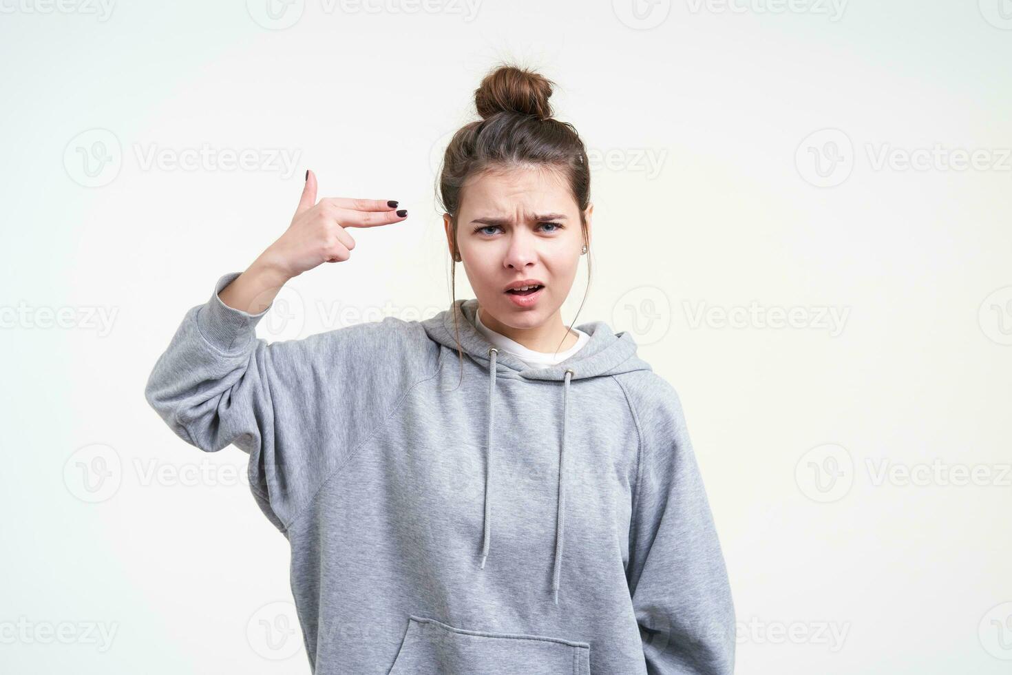 Disgruntled young brunette woman with bun hairstyle frowning her face while looking perplexedly at camera and folding gun with her fingers, isolated over white background photo