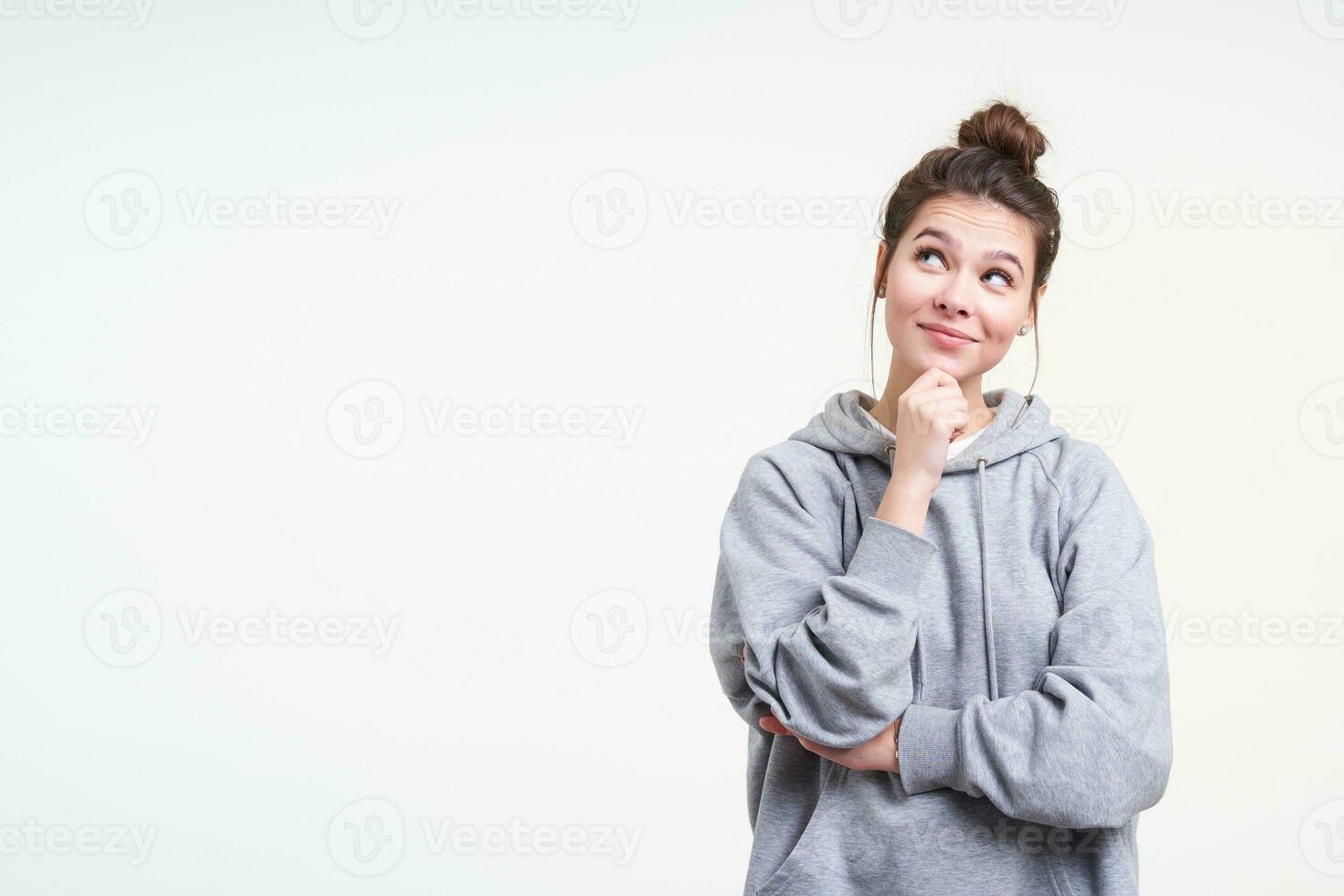 Positive young lovely brown haired female with natural makeup touching her chin while looking upwards and smiling cunningly, standing over white background photo