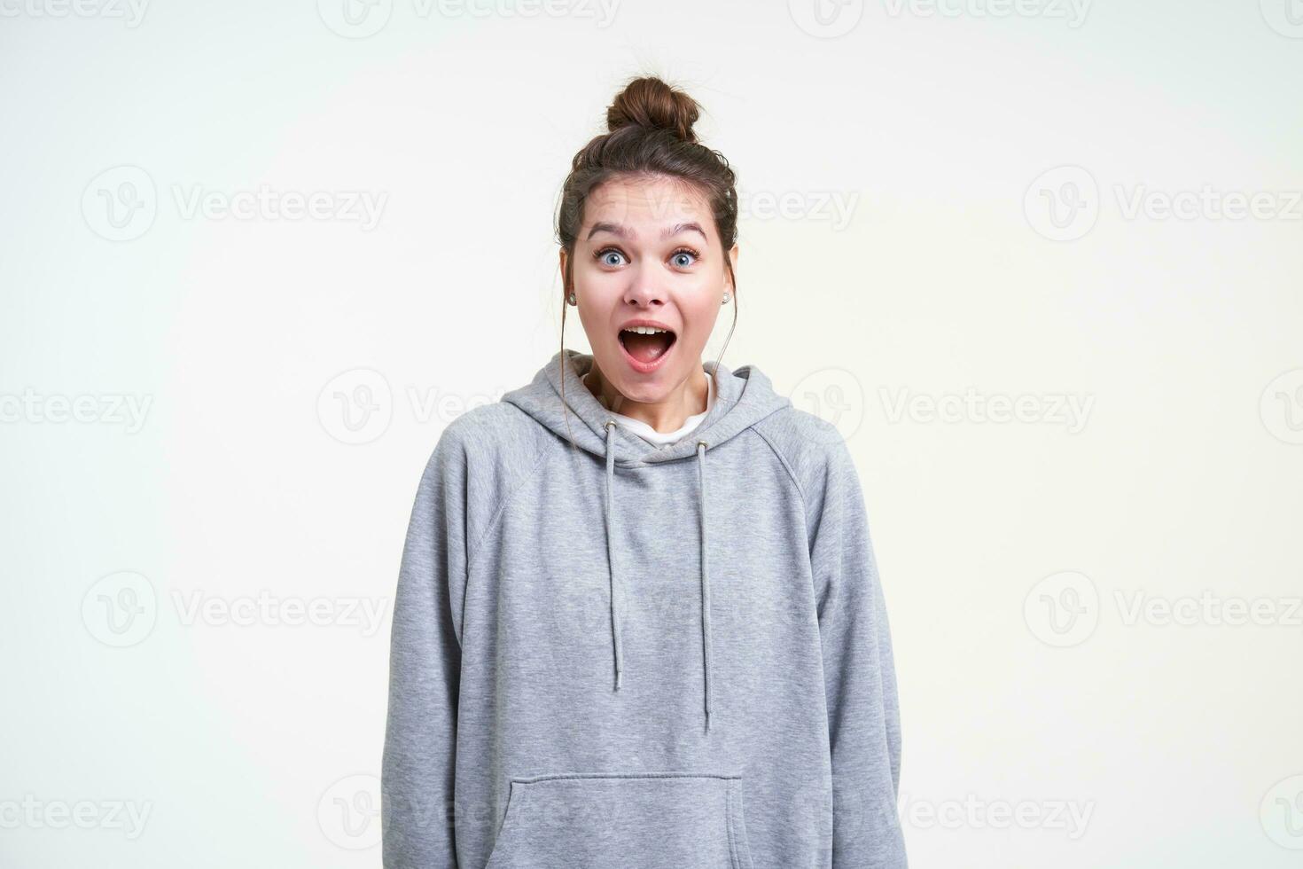 Bemused young blue-eyed brunette woman with bun casual hairstyle looking amazedly at camera with wide eyes and mouth opened, isolated over white background photo