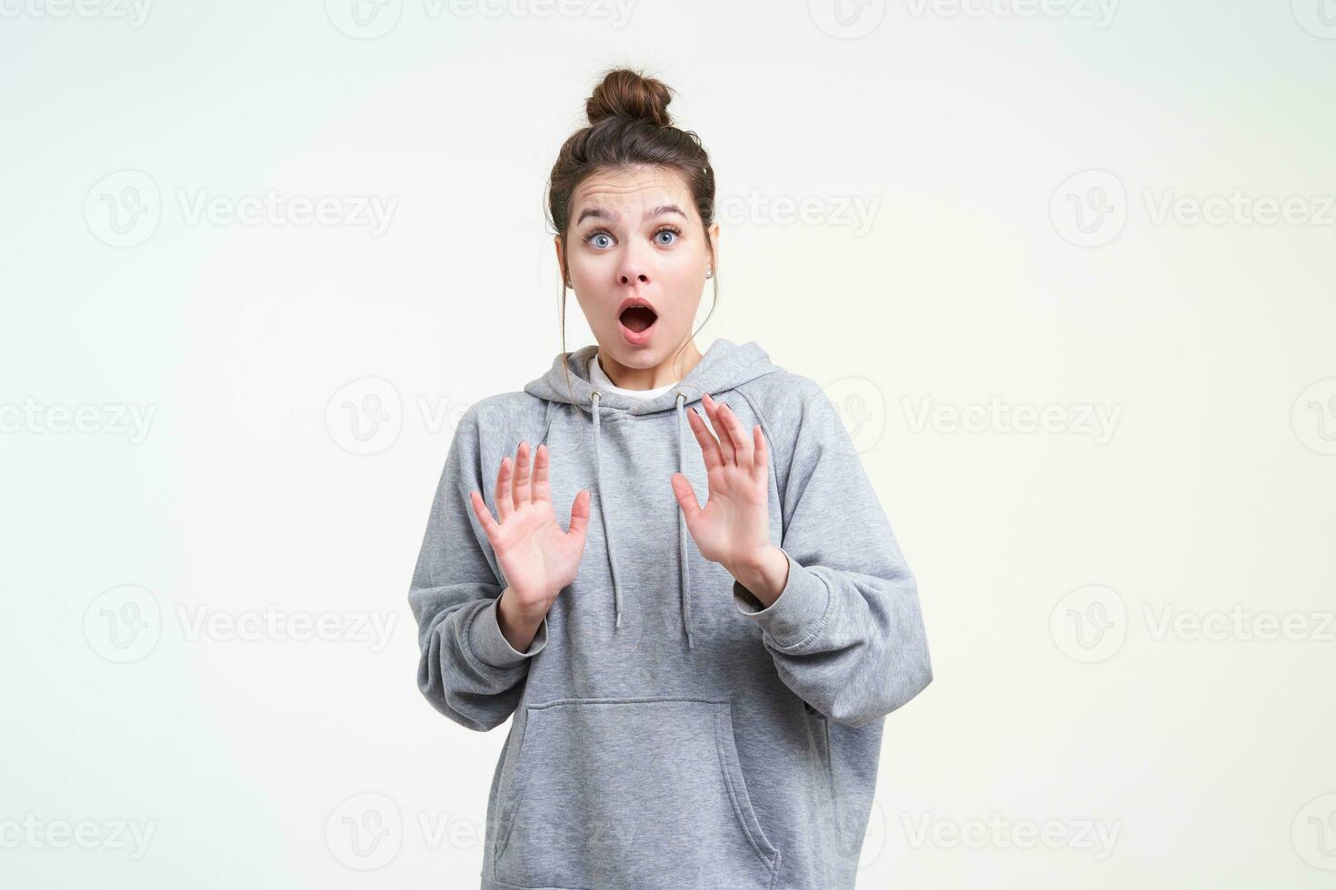 Shocked young attractive brunette female with natural makeup raising hands in protective gesture while looking dazedly at camera with wide mouth opened, isolated over white background photo