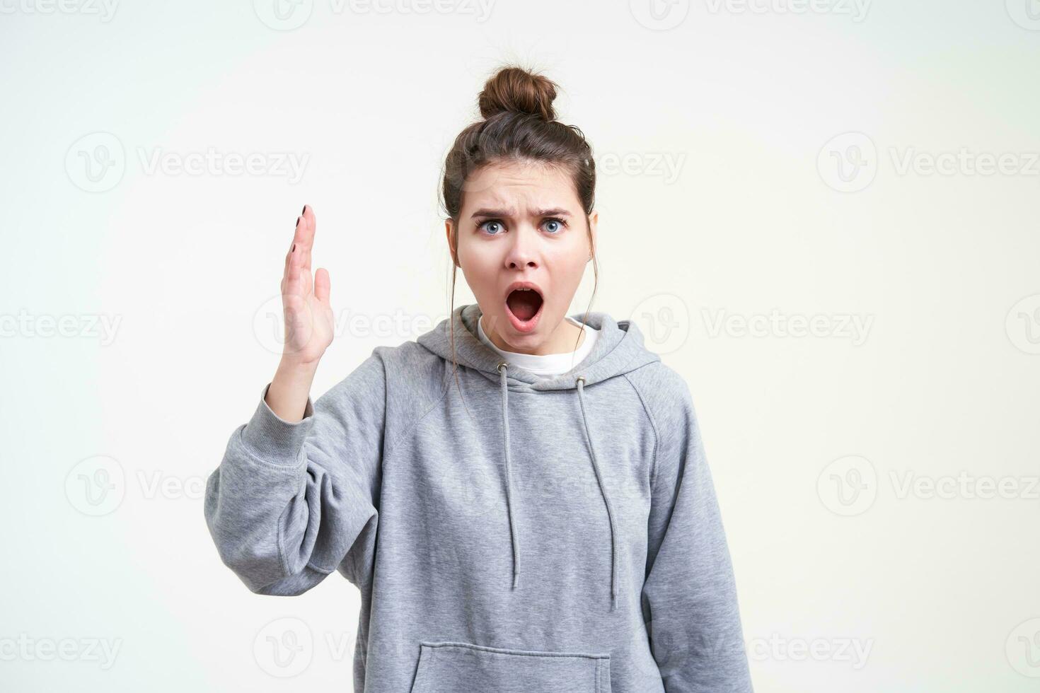 Mad young attractive long haired female with casual hairstyle raising emotionally hand while shouting angrily, standing over white background in grey hoodie photo