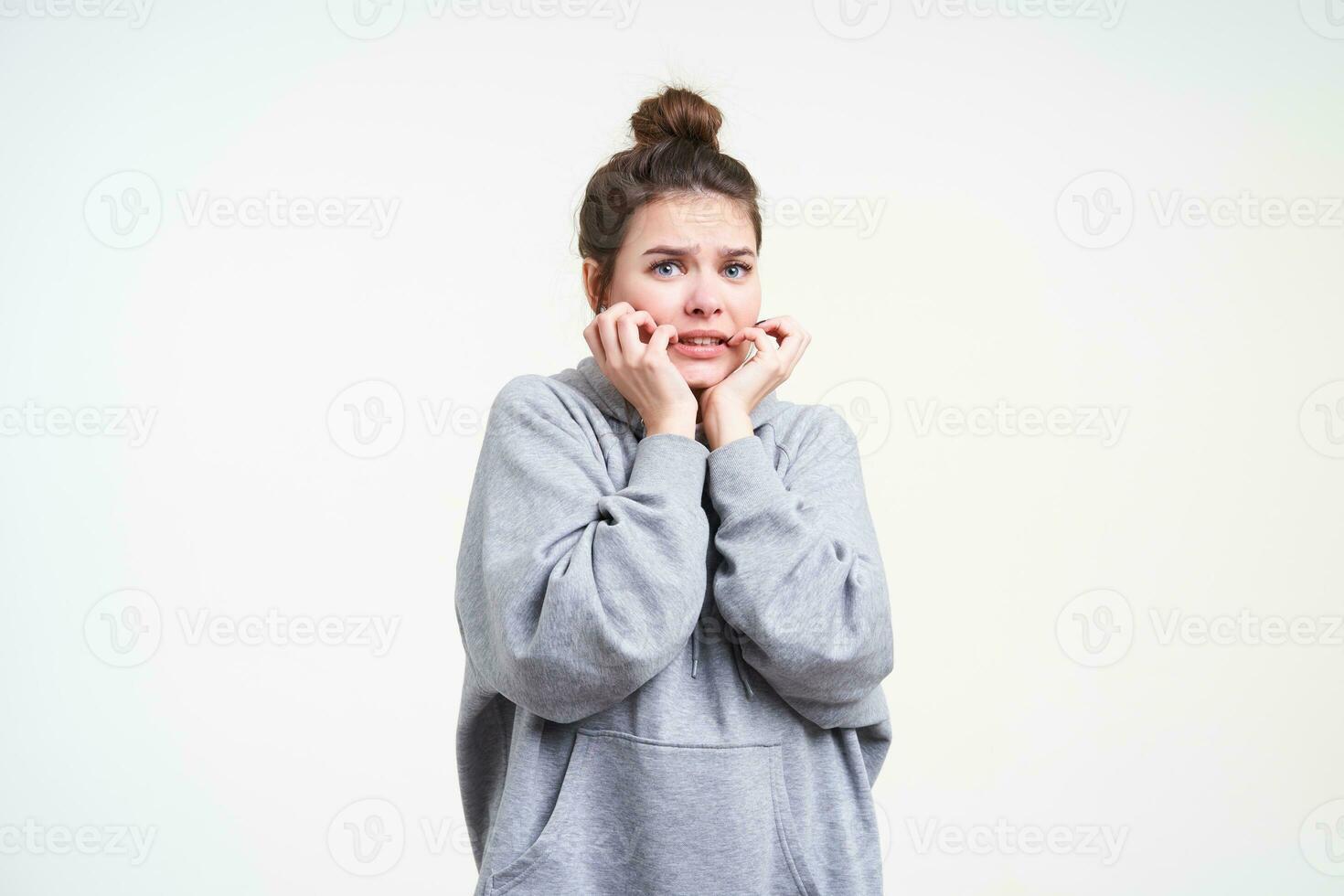Frightened young blue-eyed brown haired woman holding her face with raised hands while looking scaredly to camera, isolated over white background in grey hoodie photo