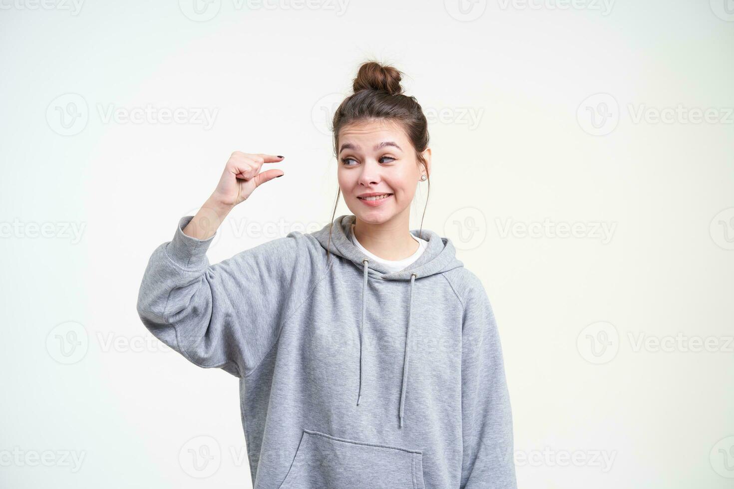 Confused young lovely long haired female with natural makeup looking ironically on her hand while showing small size with her fingers, isolated over white background photo