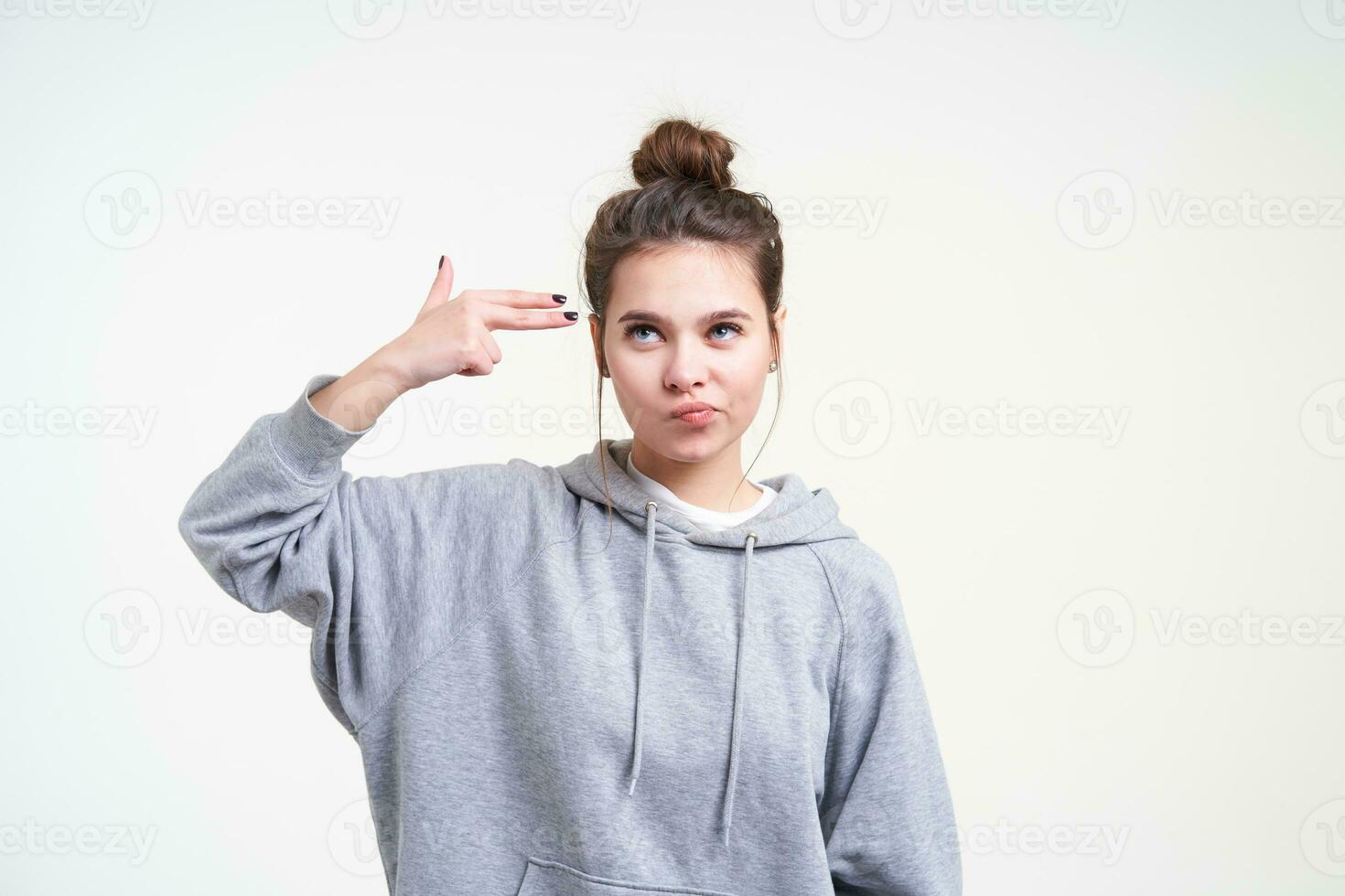 Dissatisfied young pretty long haired female wearing her brown hair in knot while posing over white background, pouting her lips while raising hand with gun gesture photo