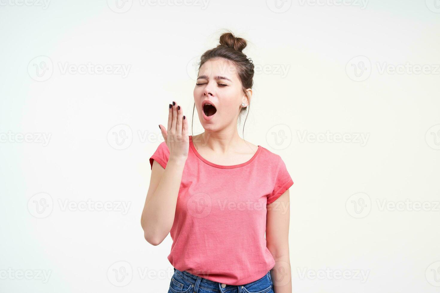 Good looking sleepy brunette young lady keeping her eyes closed while yawning and going to cover her mouth with raised hand, isolated over white background photo