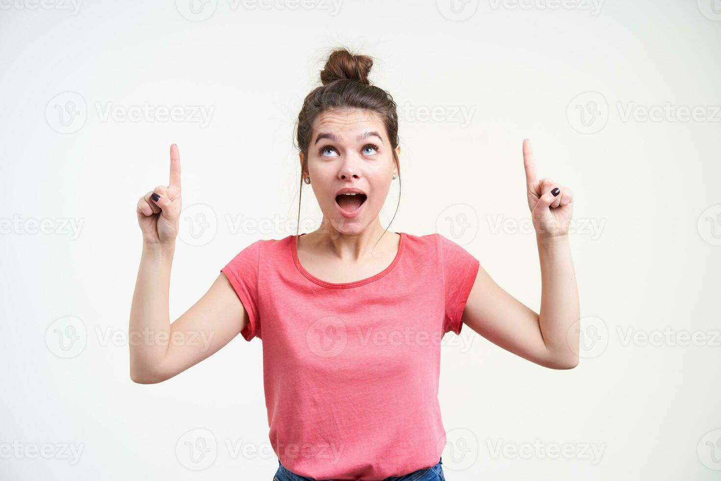 Agitated young lovely brown haired female keeping her mouth opened while pointing excitedly upwards with index fingers, isolated over white background photo