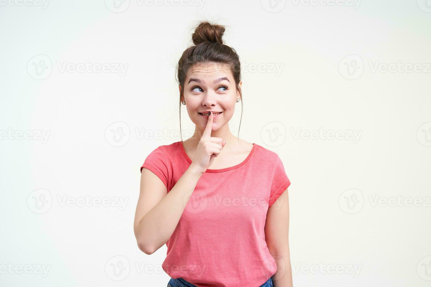 Glad young lovely brown haired female with natural makeup looking positively aside while keeping forefinger on her nouth, standing over white background photo