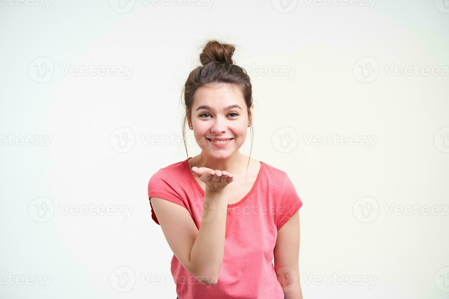 Indoor photo of young blue-eyed pretty brunette female holding raised hand and smiling cheerfully while looking gladly at camera, standing over white background