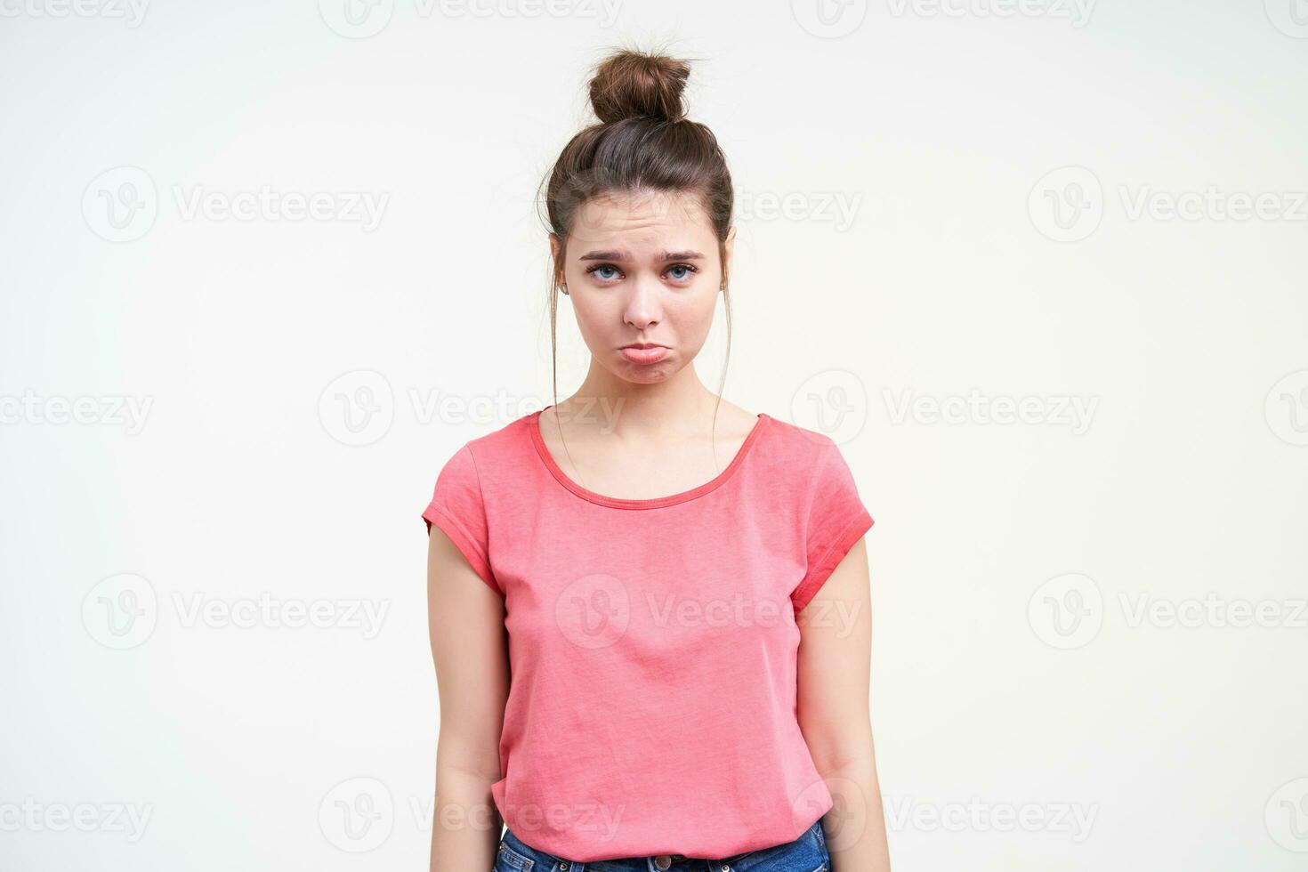 Offended young brown haired woman with natural makeup pouting her lips while looking sadly at camera, keeping hands along body while posing over white background photo