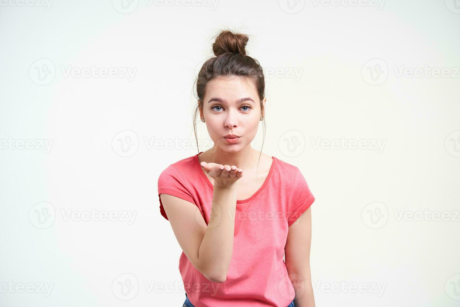 Studio photo of young attractive brown haired lady keeping palm raised while blowing air kiss at camera, standing over white background in pink t-shirt