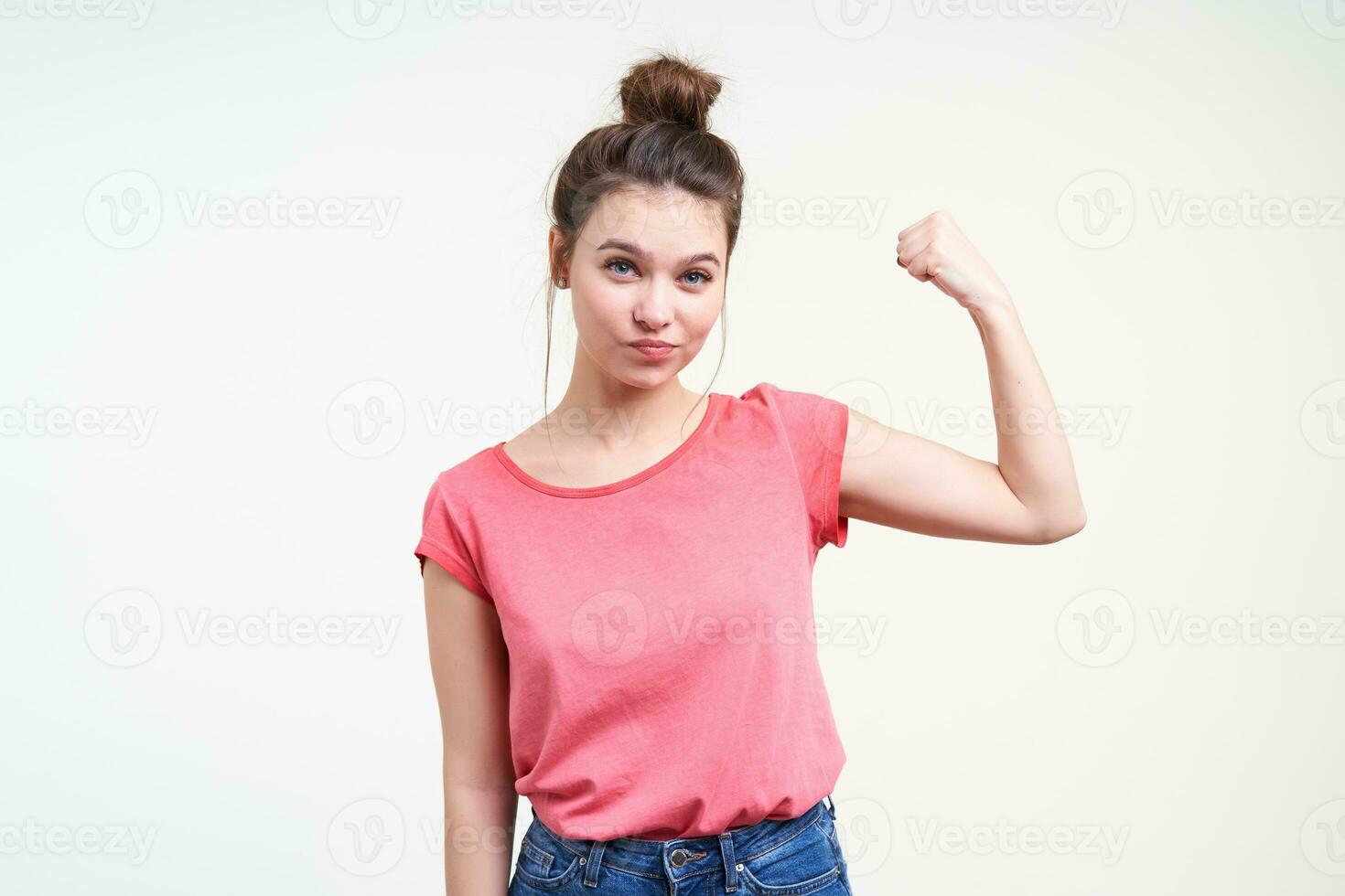 Studio photo of young charming brown haired woman with bun hairstyle looking attentively at camera while showing her power with raised hand, isolated over white background