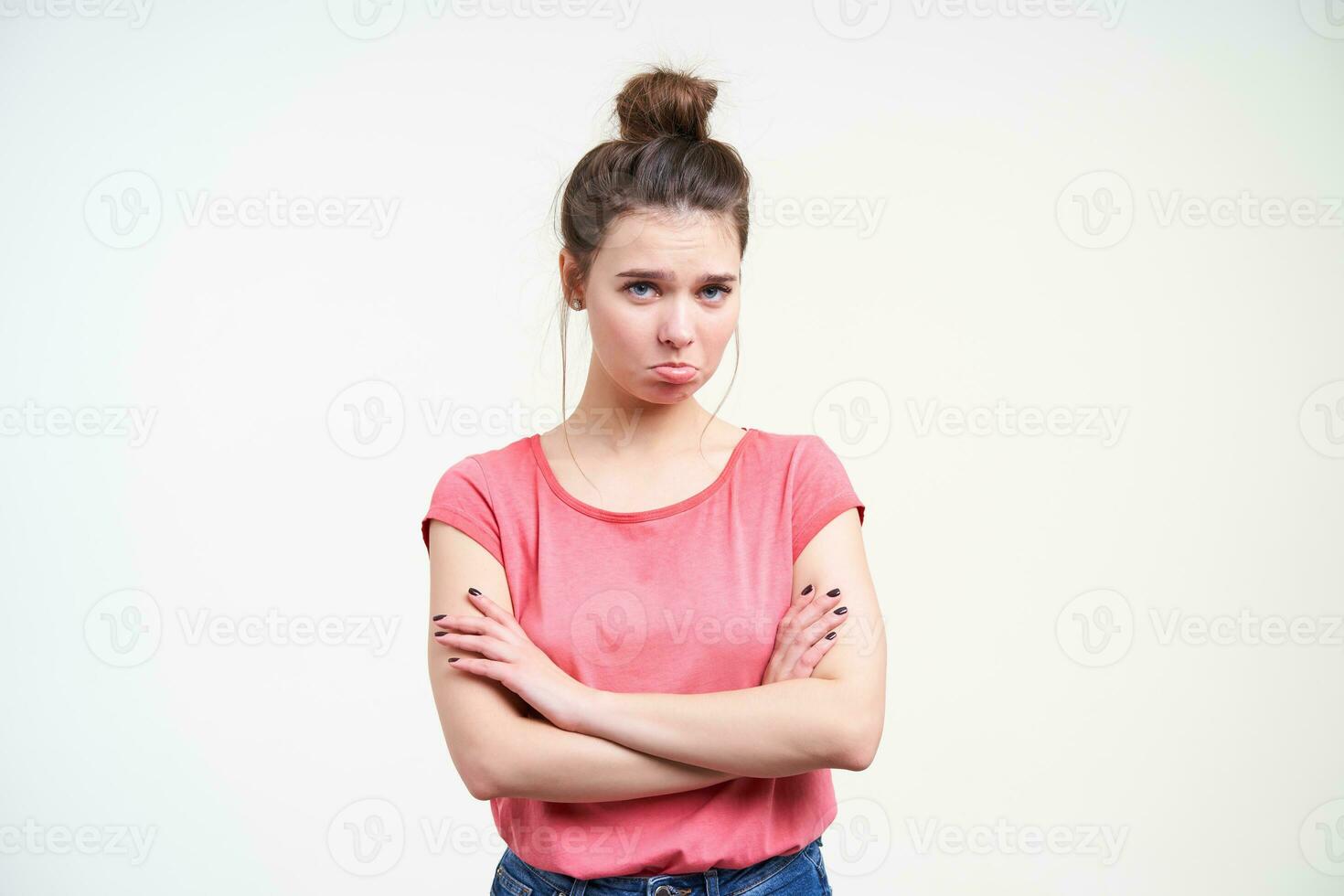 Unhappy young pretty brunette female with bun hairstyle keeping hands crossed on her chest while looking resentfully at camera, being isolated over white background photo