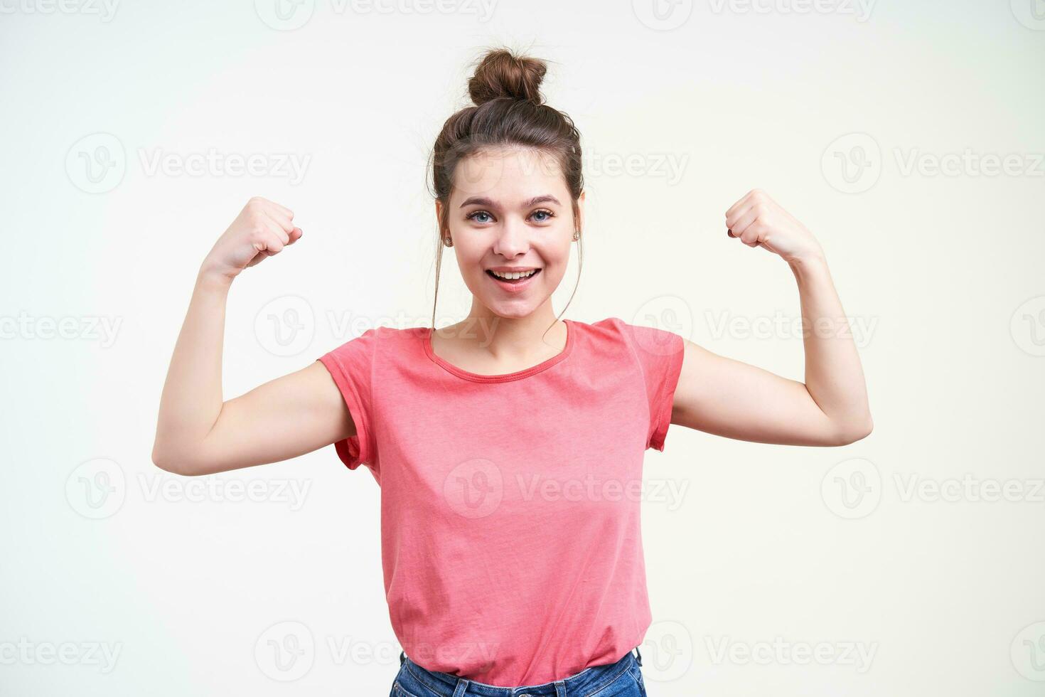 Cheerful young attractive brown haired woman with natural makeup raising her hands while showing biceps and smiling gladly at camera, isolated over white background photo