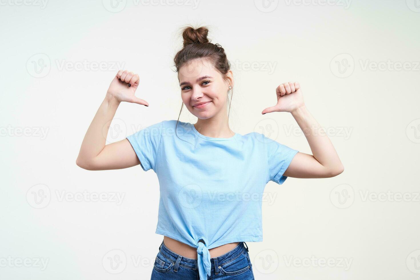 Pleased young lovely brunette female dressed in casual clothes smiling positively while thumbing on herself with raised hands, isolated over white background photo