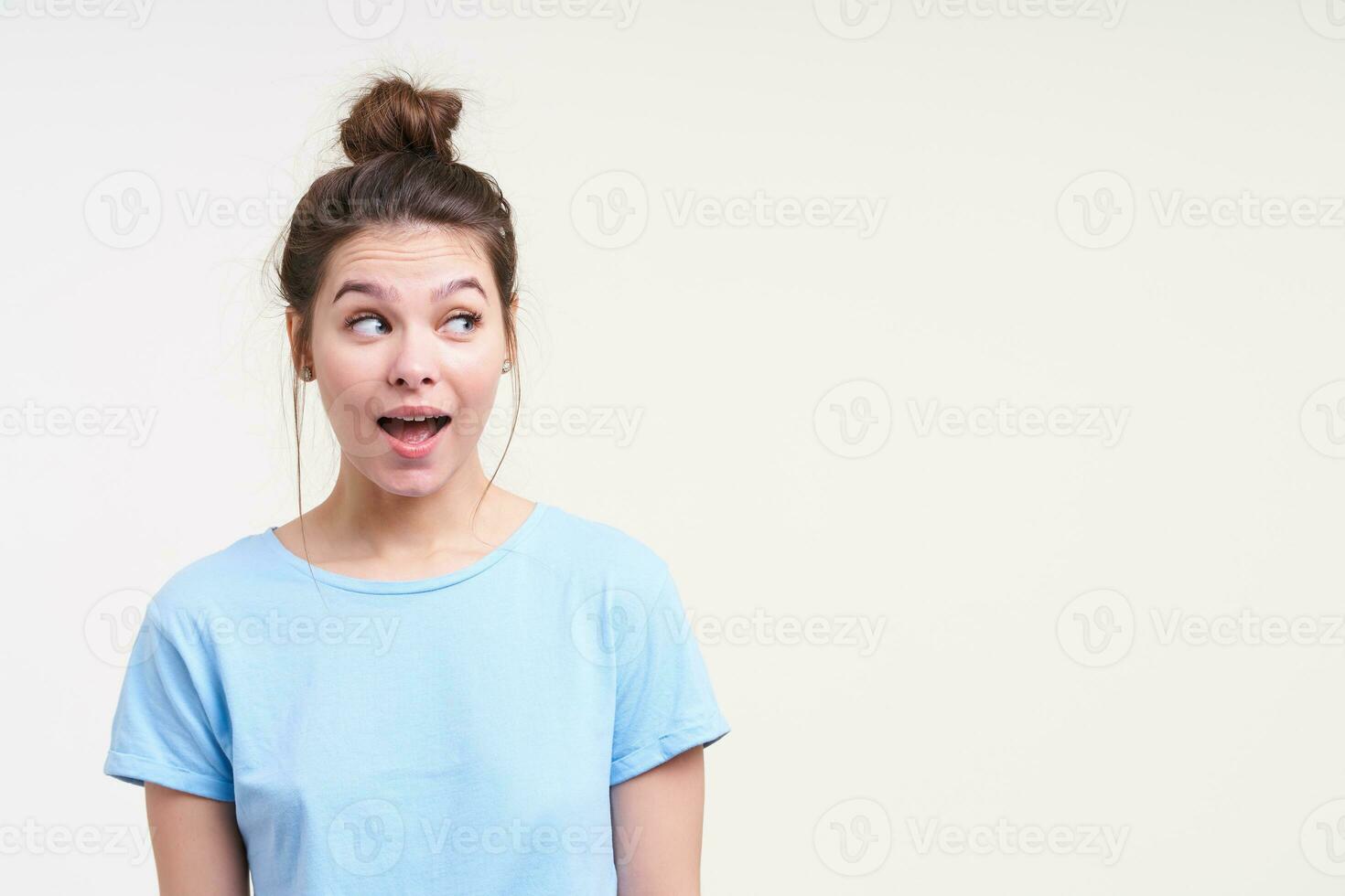 Portrait of young blue-eyed dark haired female raising surprisedly eyebrows while looking excitedly aside with wide mouth opened, isolated over white background photo