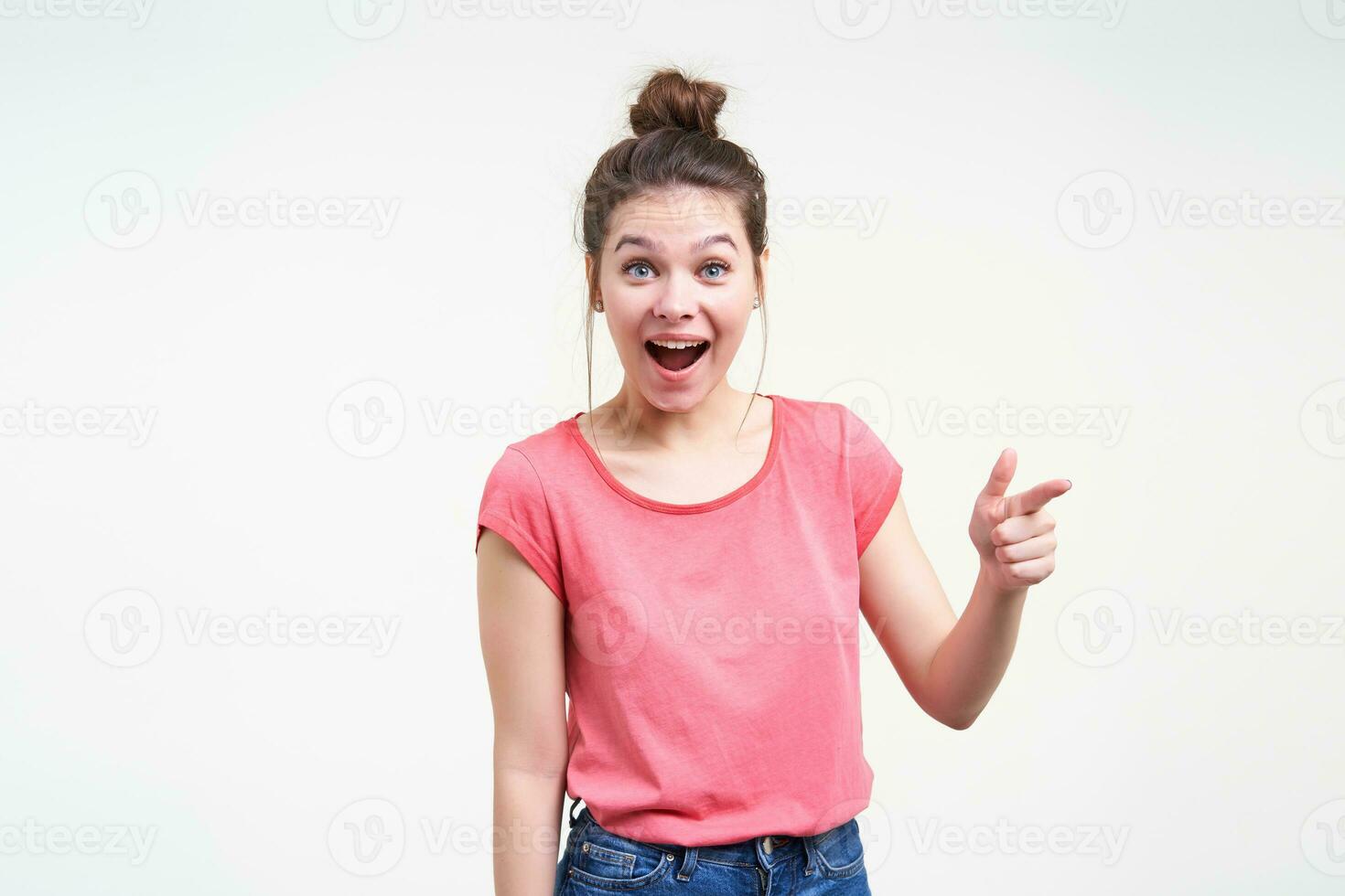 Excited young pretty dark haired female with natural makeup looking amazedly at camera with wide mouth opened while showing aside with forefinger, isolated over white background photo